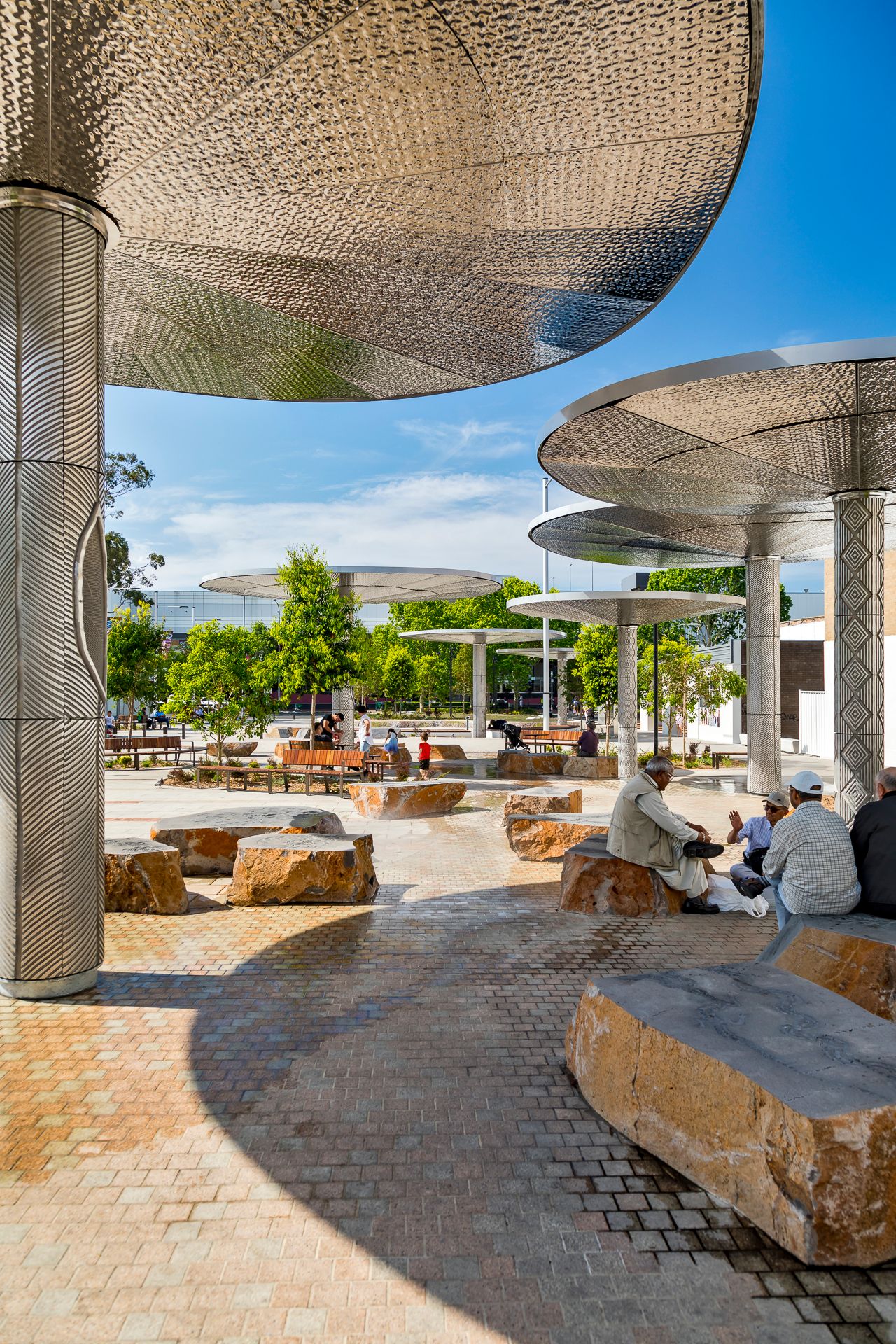 People relax on sandstone boulder seating beneath large perforated metal canopies in a sunny paved urban plaza.