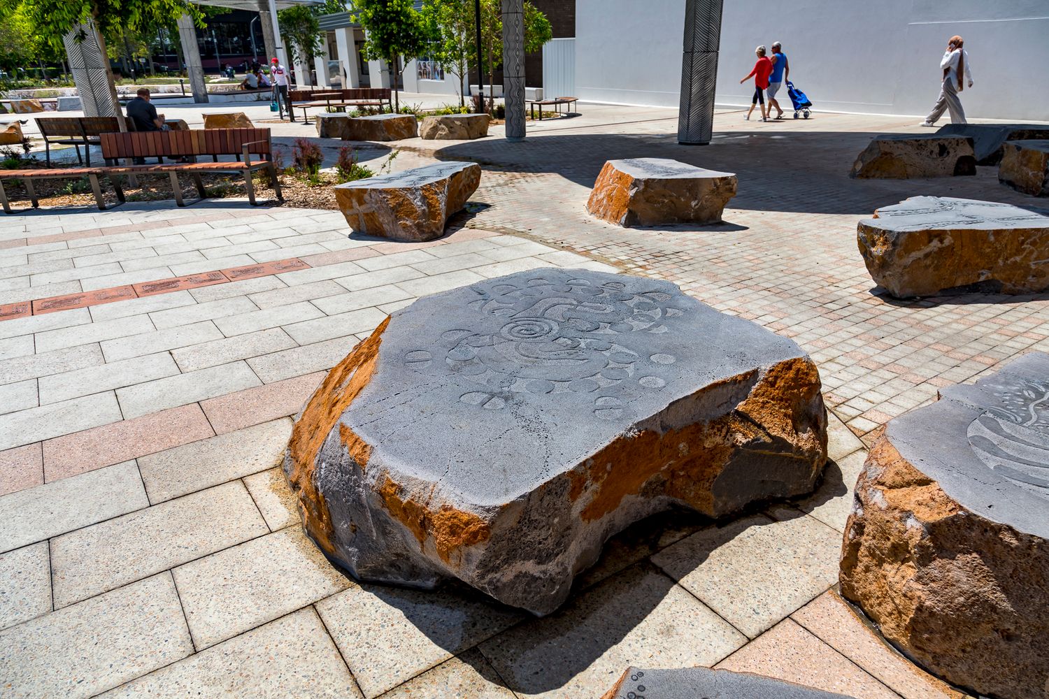 Carved stone boulders set into a paved plaza with timber benches and pedestrians walking in the background.