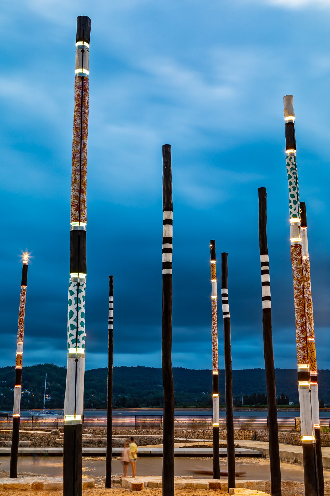 Illuminated Aboriginal-patterned poles glow at dusk against a deep blue sky with two people walking below near the water.