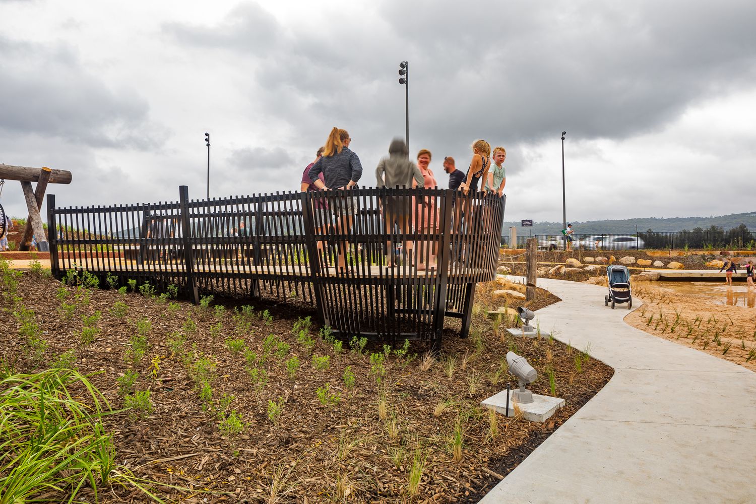 Visitors gather on an elevated wooden viewing deck beside landscaped garden beds at a waterfront public park on a cloudy day.