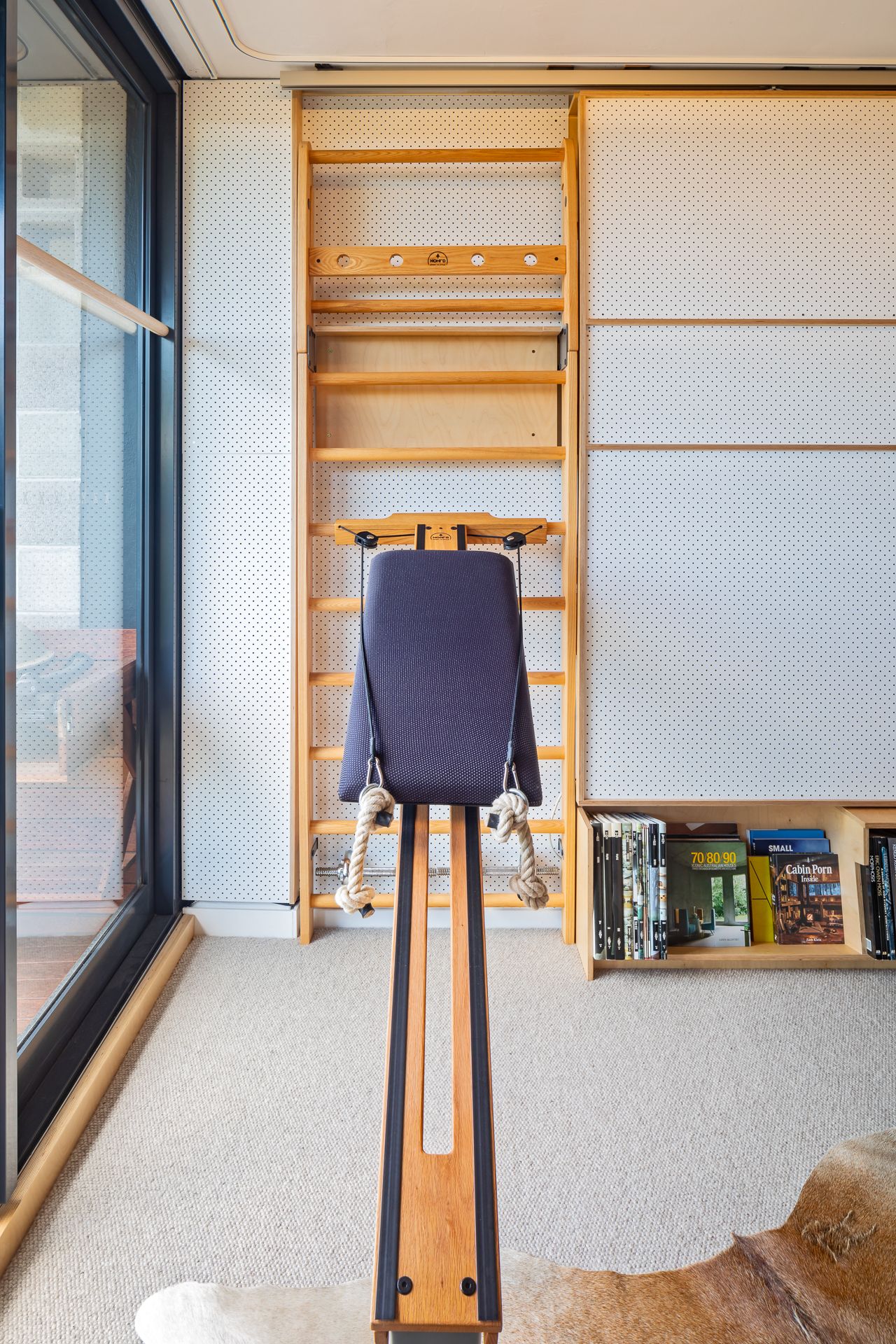 Home gym corner with a wooden rowing machine facing wall bars, white panels and a sunlit glass door.