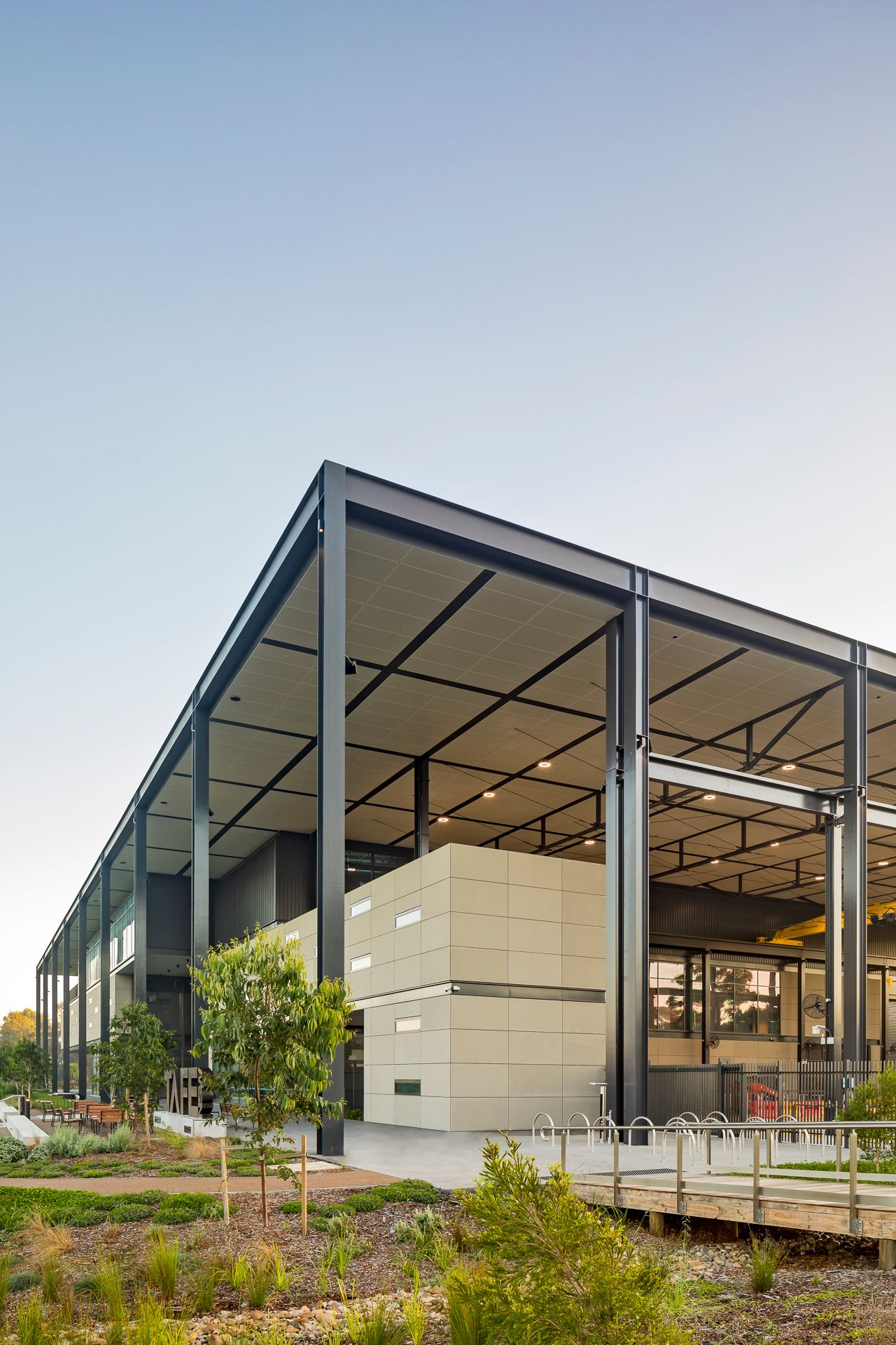 Corner of a modern steel-framed building with beige cladding panels, deep roof canopy, native plantings and a timber walkway.