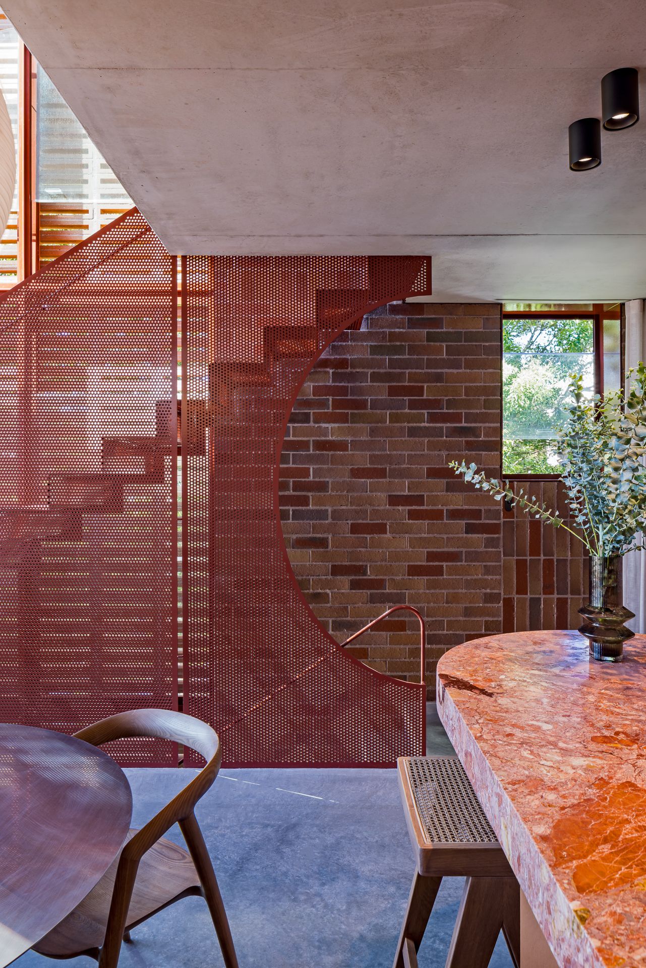 Dining area featuring a red perforated stair screen, curved brick wall, pink marble table and polished concrete ceiling.