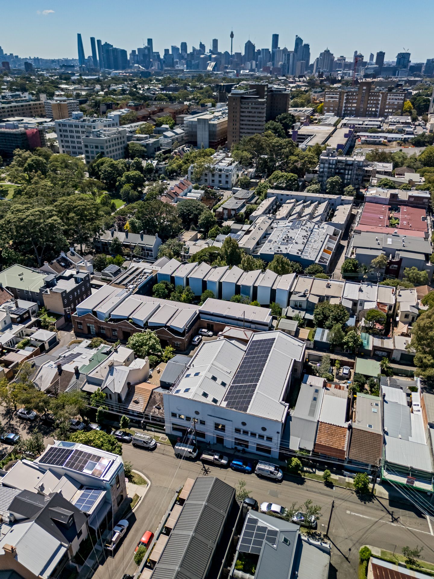 Aerial view of inner-city suburban rooftops and tree-lined streets with a distant city skyline under a clear blue sky.