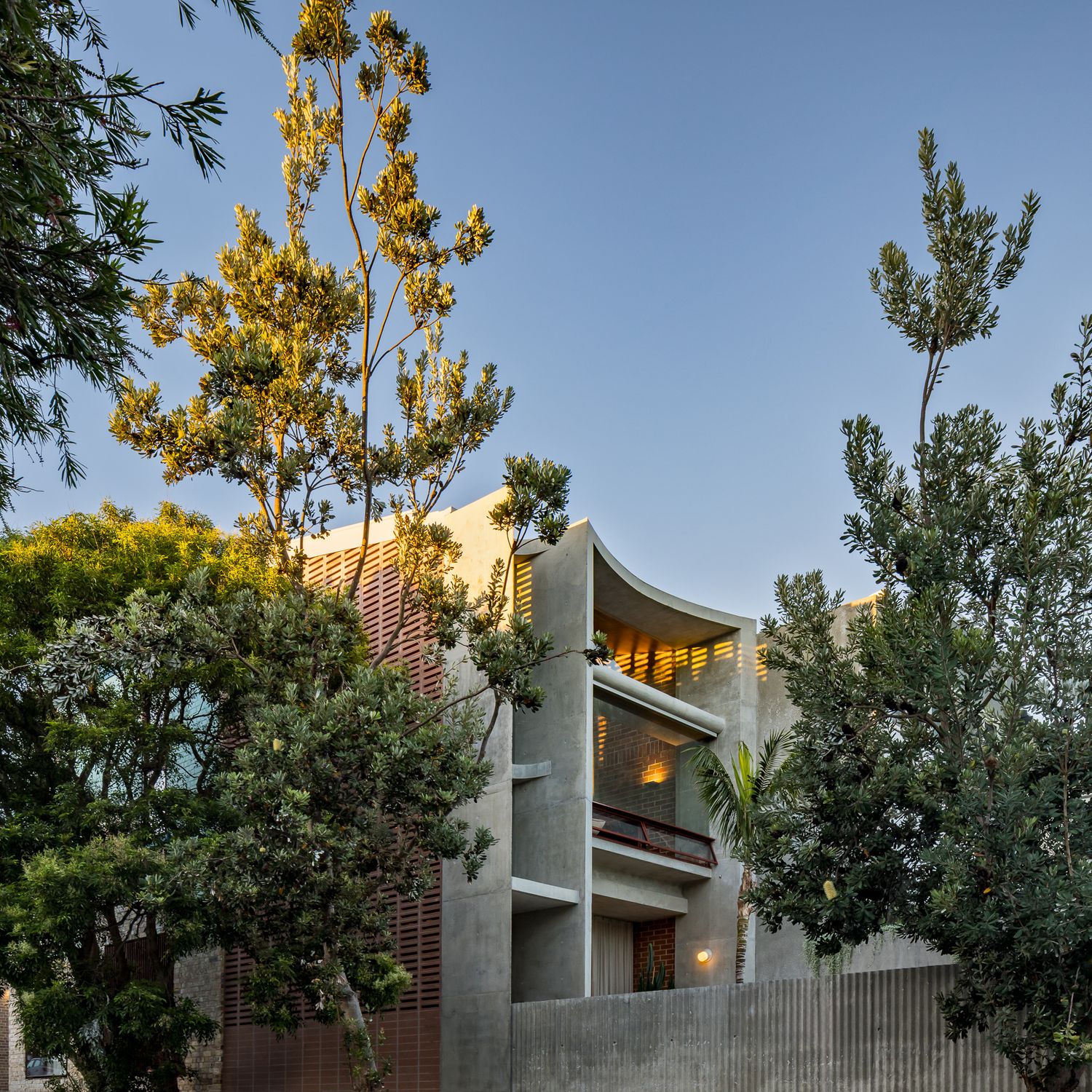 Contemporary concrete apartment building with curved balconies and timber screens, partly framed by lush green trees at dusk.