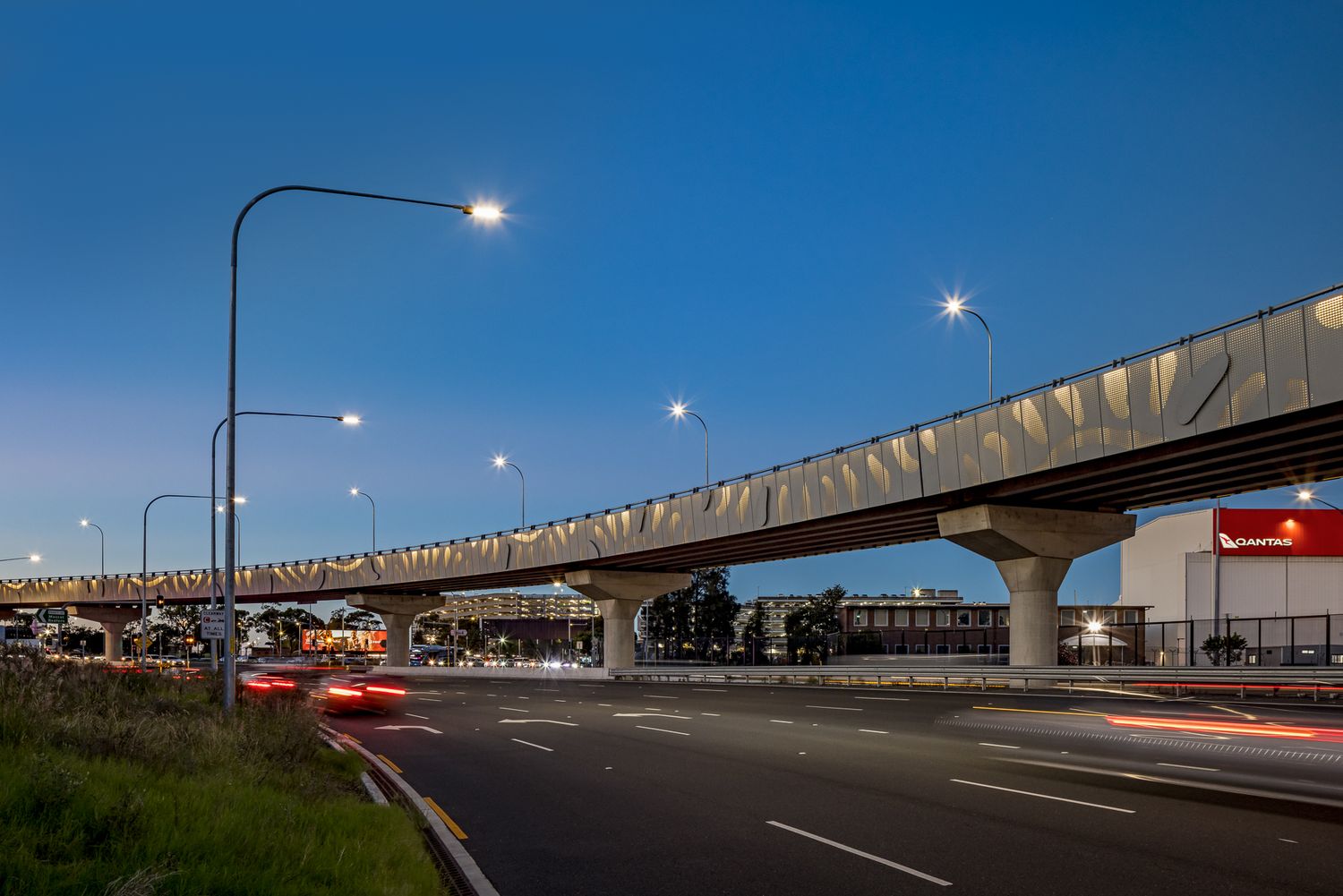 Curving elevated railway on concrete piers rises above a quiet road at dusk, with glowing street lights and blurred red vehicle trails.