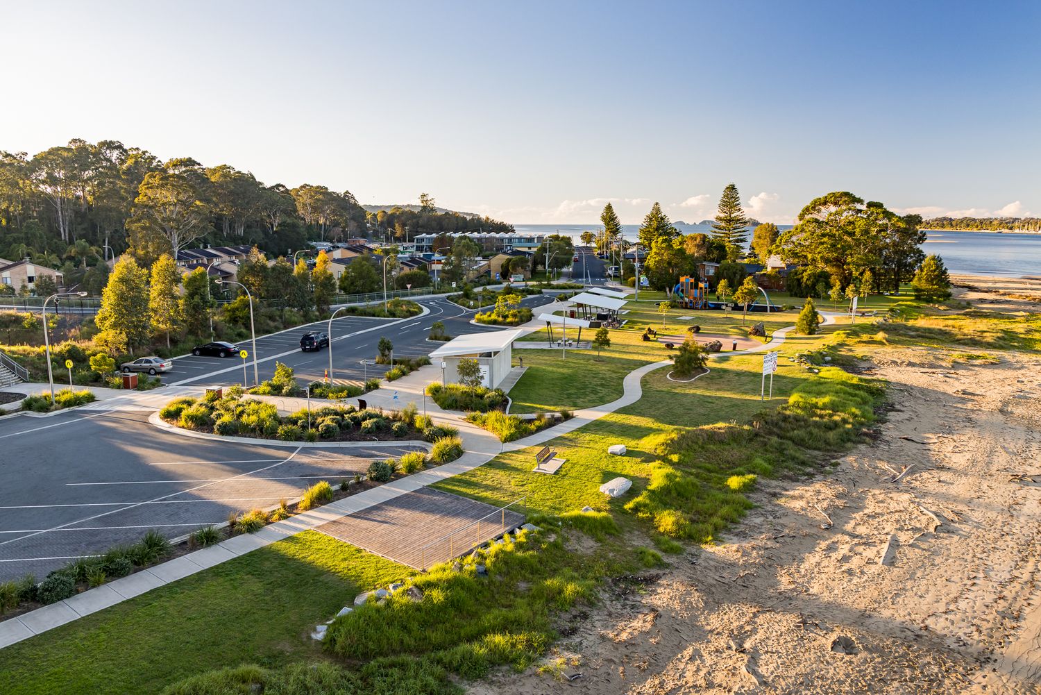 Aerial view of a waterfront park with winding paths, car park, trees, sandy beach and nearby homes beside a calm bay at sunset.