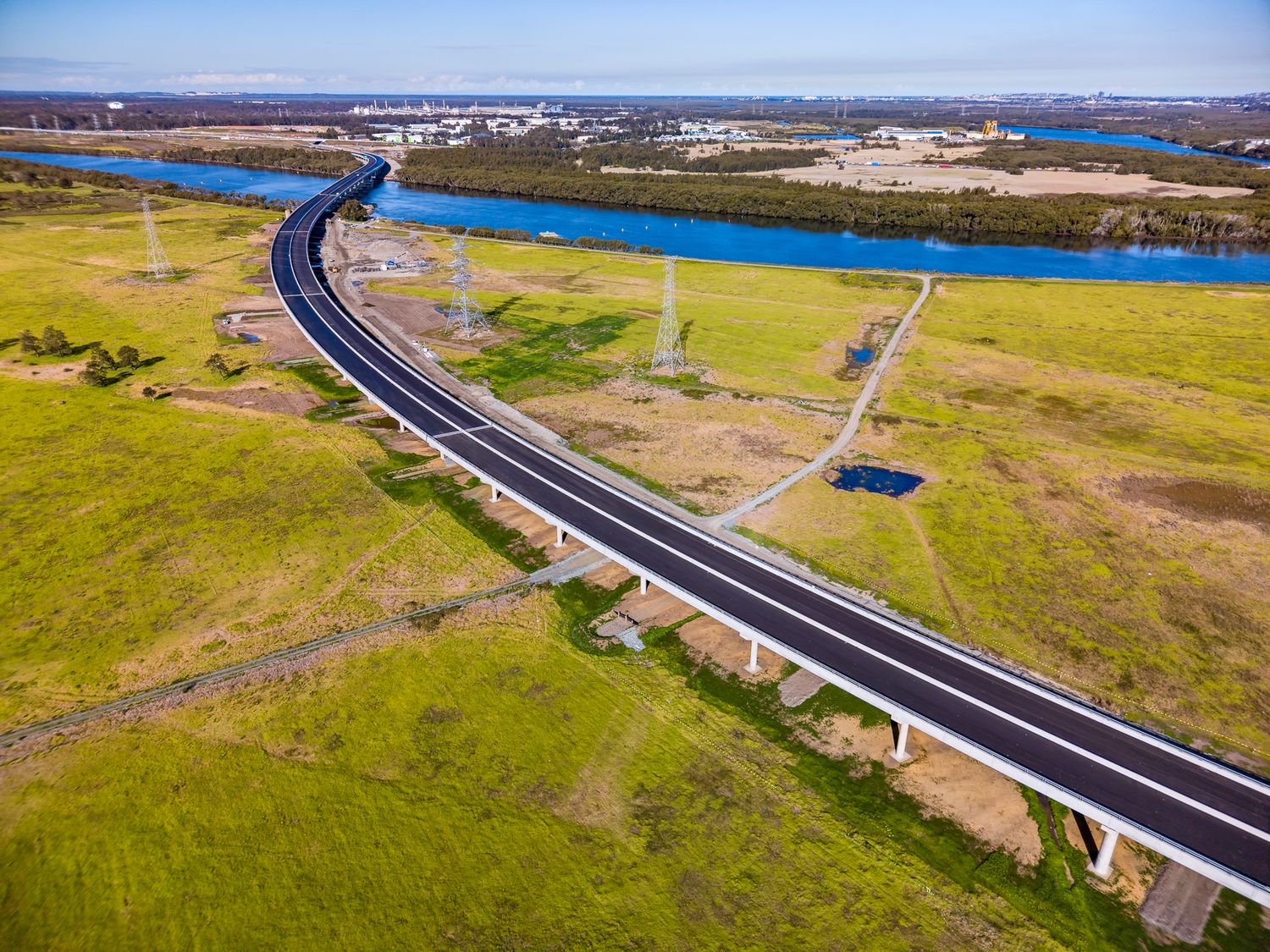 Aerial view of a curved new highway and bridge sweeping across green grassland toward a wide blue river.