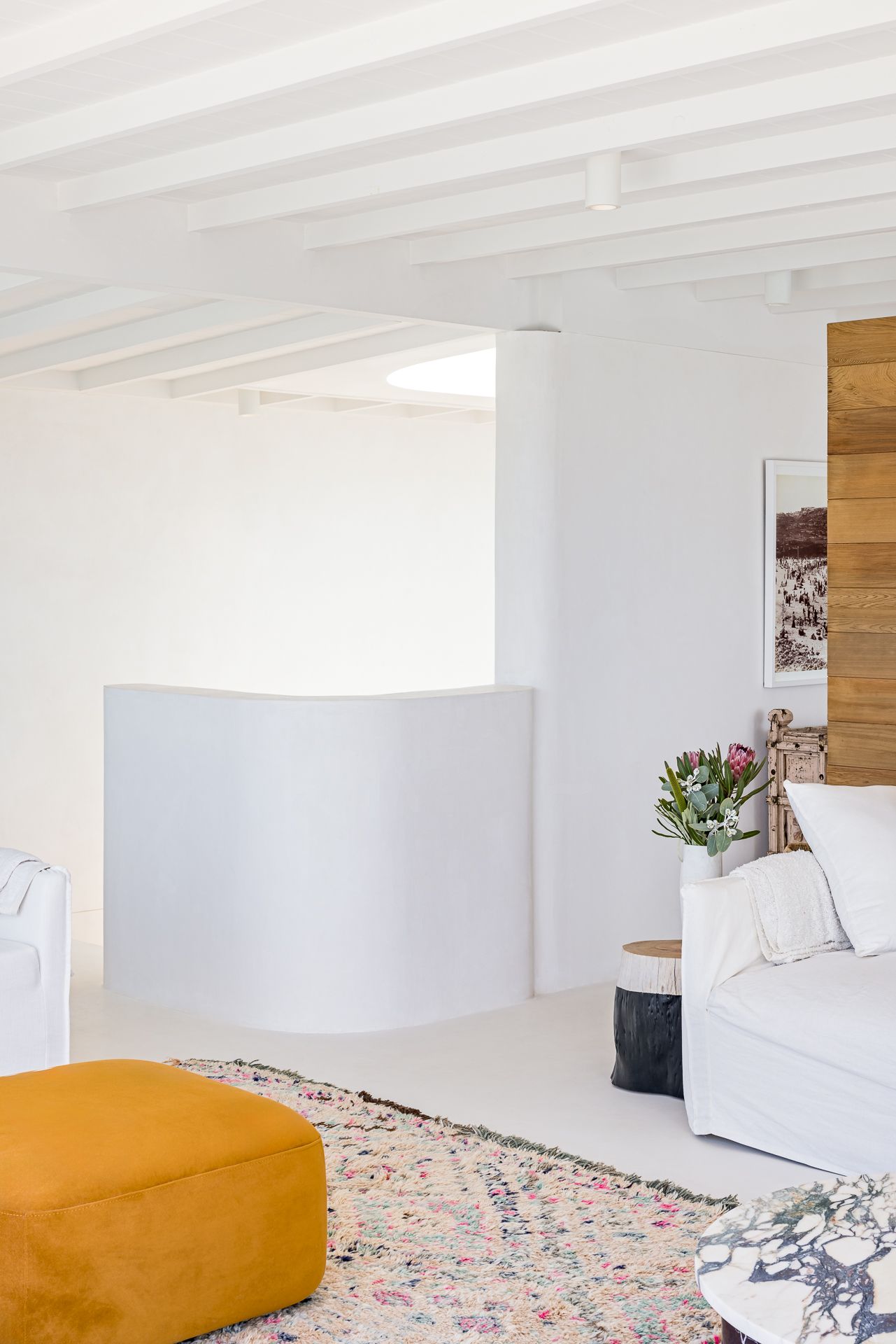 Bright white living space with exposed beams, mustard ottoman, boho patterned rug, slipcovered sofa and pink flowers.