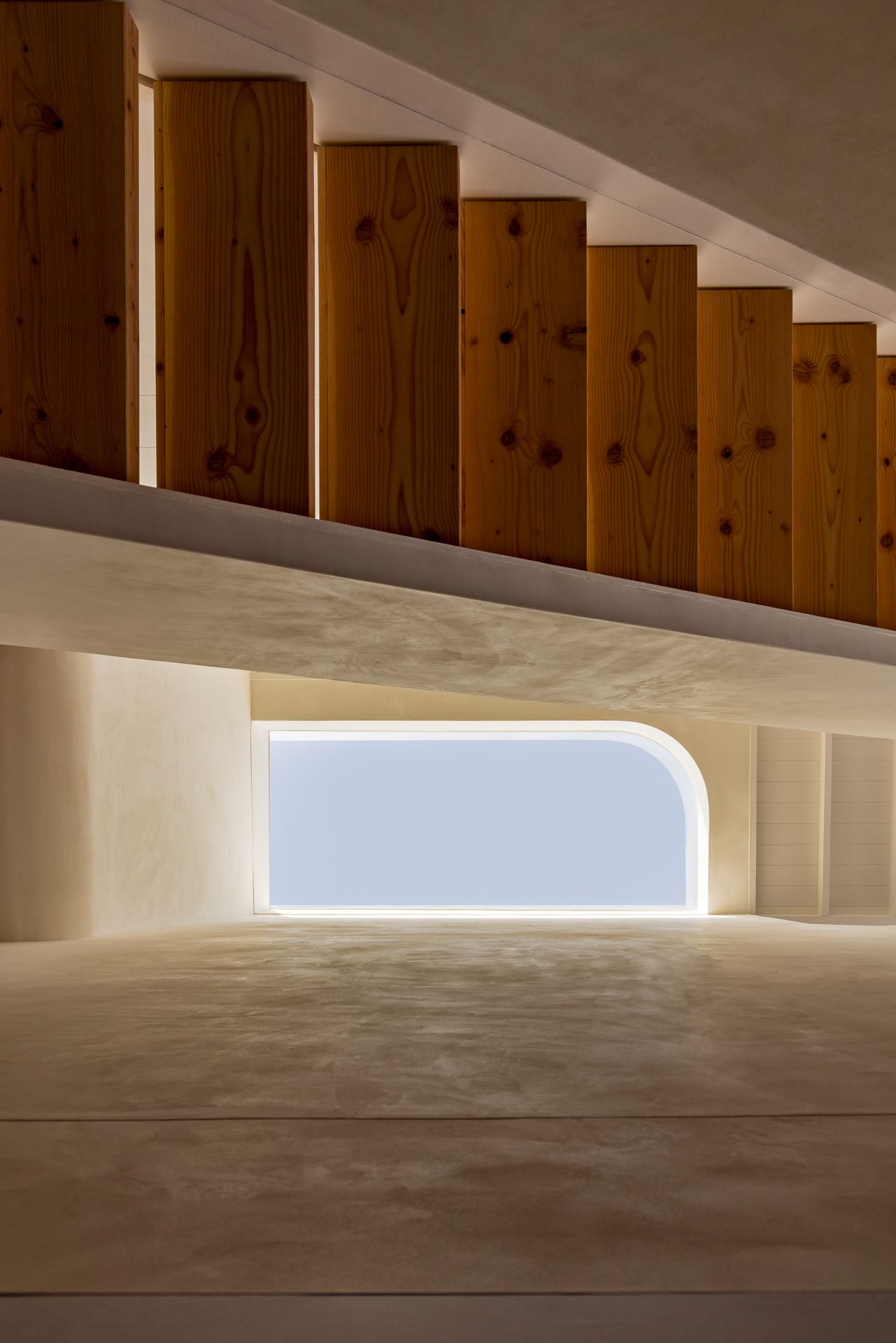 Minimalist interior with warm pine wood slats above a curved glowing window and smooth pale plaster walls.