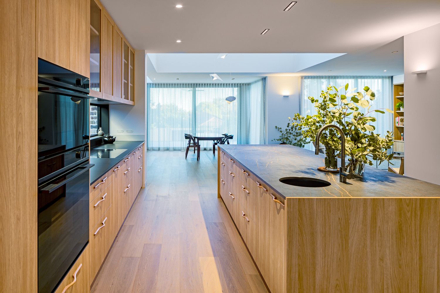 Modern timber kitchen with a long island, undermount sink, black built-in ovens and a dining area leading to a sunlit garden.
