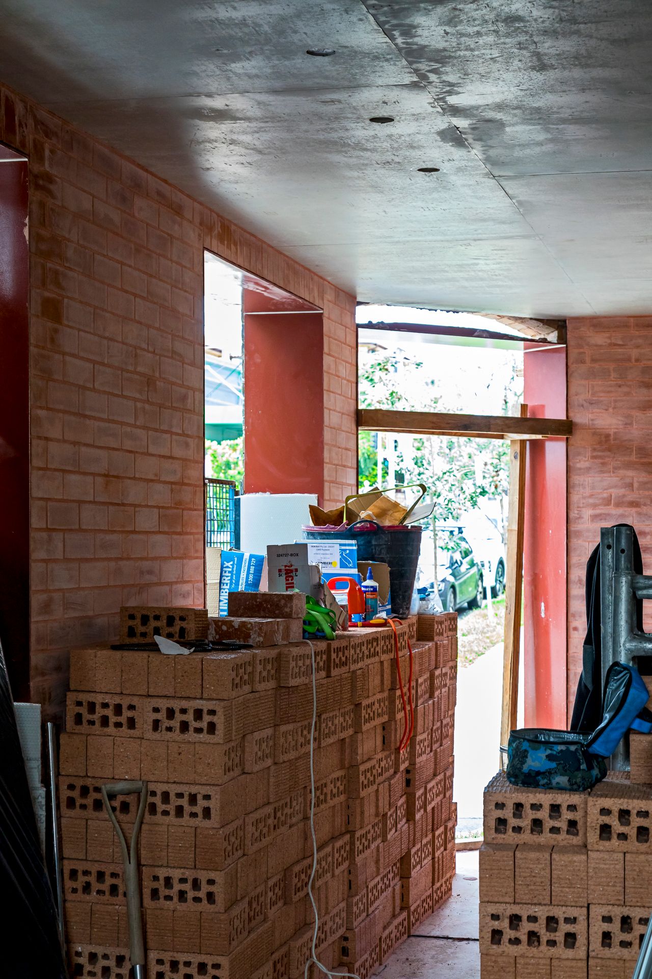 Stacks of perforated bricks and tools beneath a concrete ceiling in a half-built room with red brick walls and open doorways.