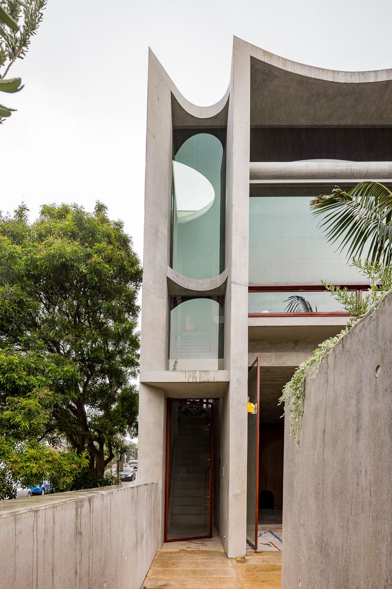 Sculptural concrete facade with curved arched windows, a timber entry door and leafy tropical plants beside a concrete wall.