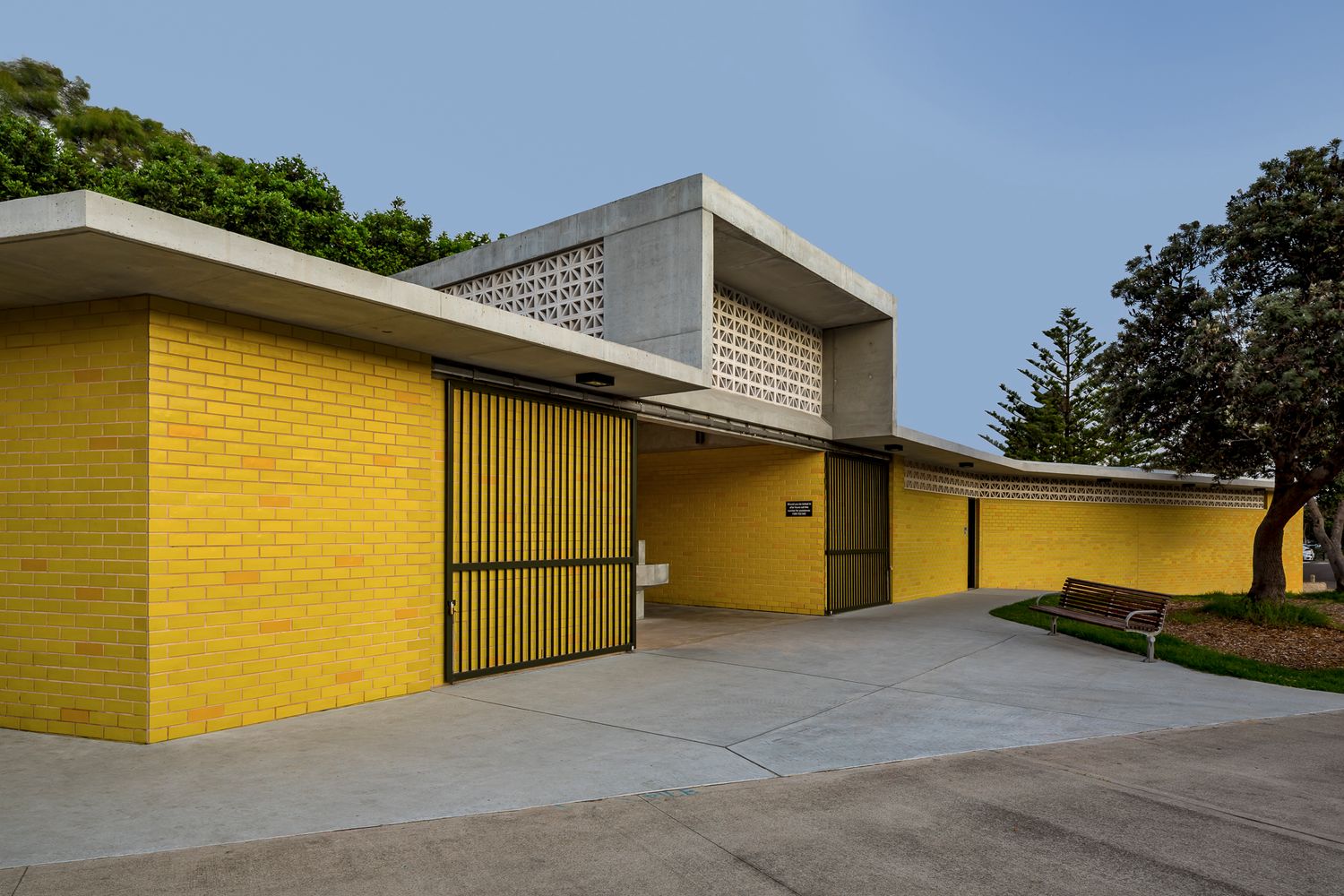 Yellow brick amenities building with slatted timber gates, a raised concrete roof volume, a curved path and a park bench under mature trees.