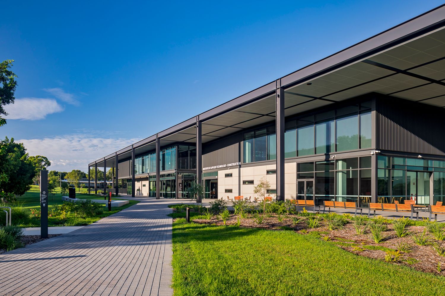 Paved pathway leading to a modern TAFE building with glazed walls, steel columns and landscaped garden beds in warm sunlight.