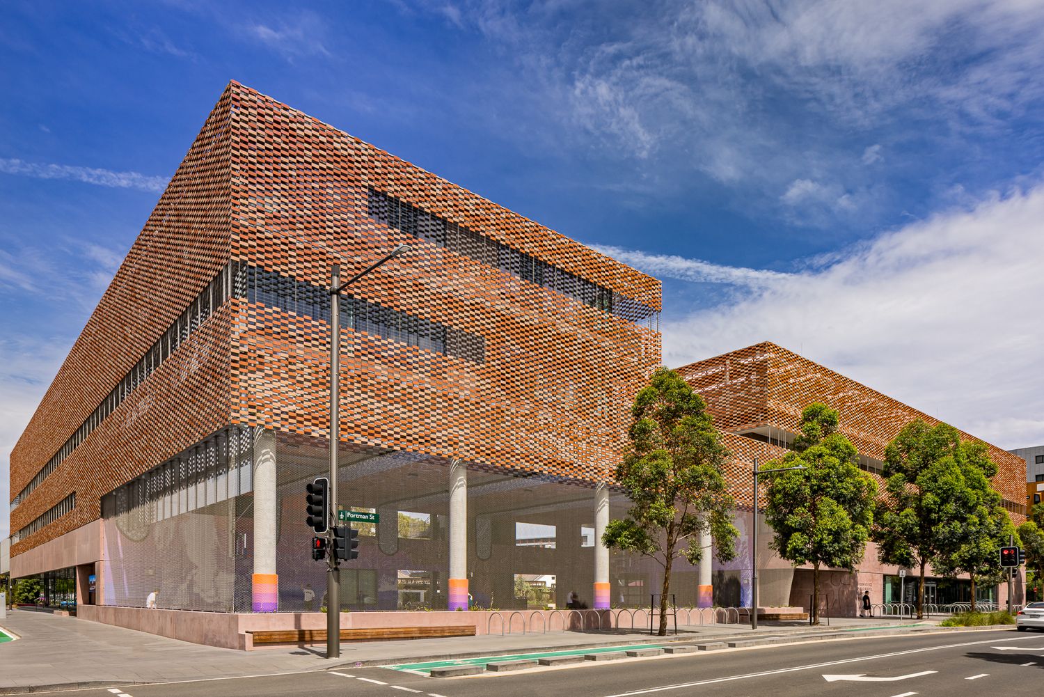 Modern brick-clad building with perforated facade and white columns beside a city street under a blue sky with trees.