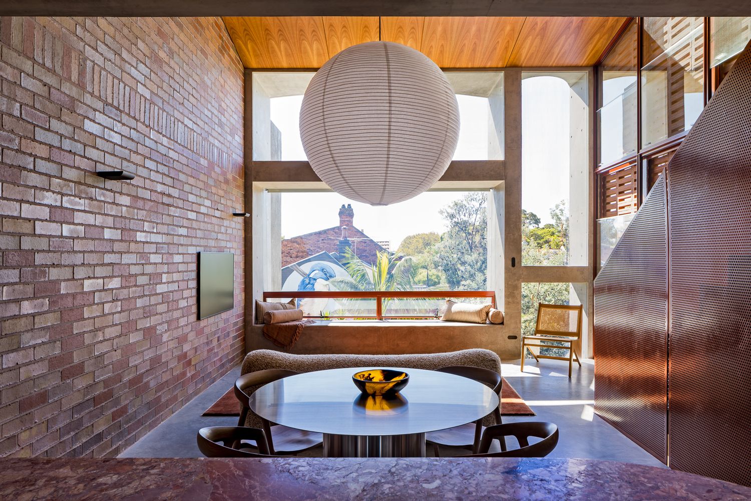 Modern dining area with a round table and pendant lamp beside a brick wall and a window seat overlooking leafy treetops.