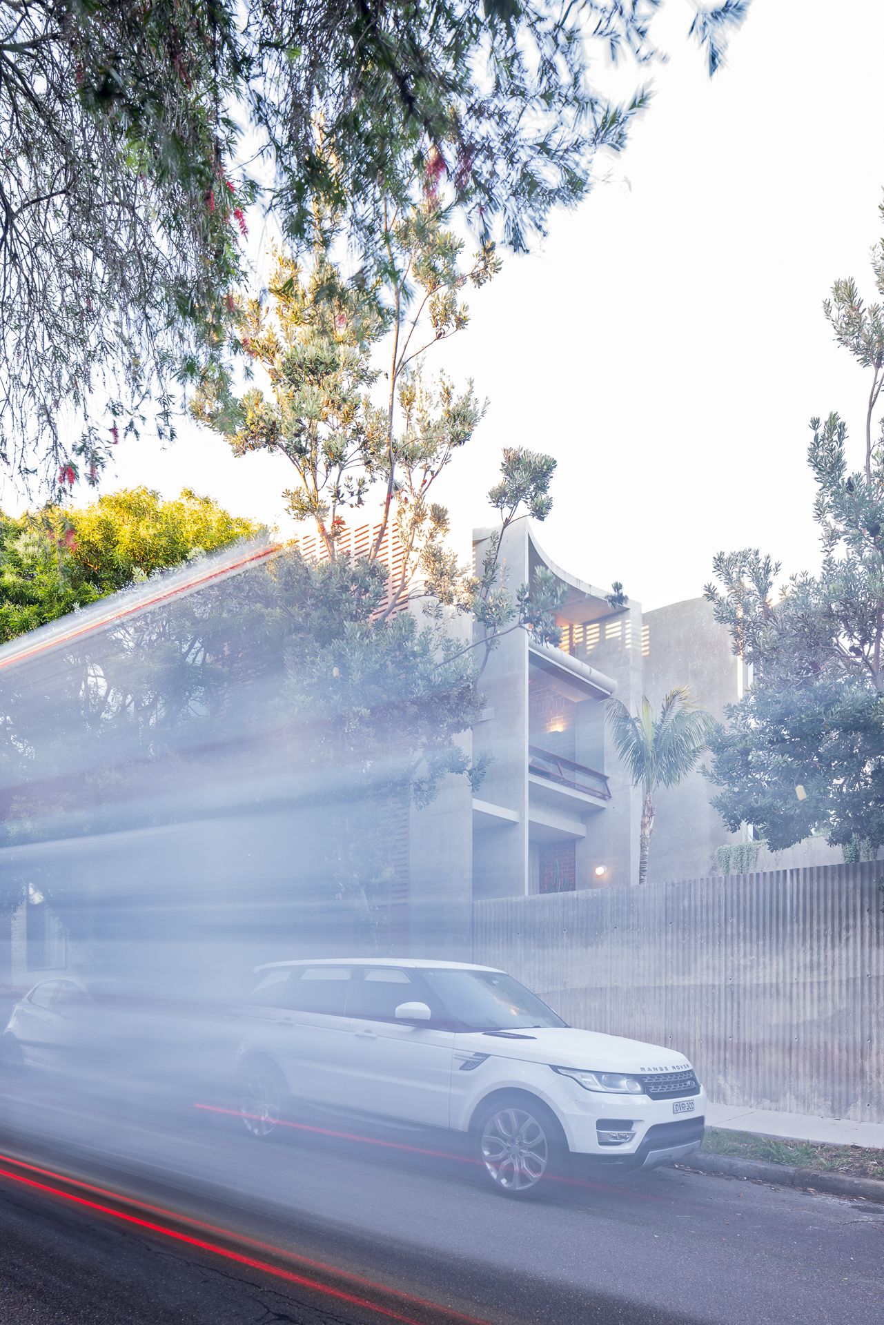 White Range Rover passes a modern concrete townhouse behind a slatted fence, with motion-blurred light streaks and trees in the foreground.