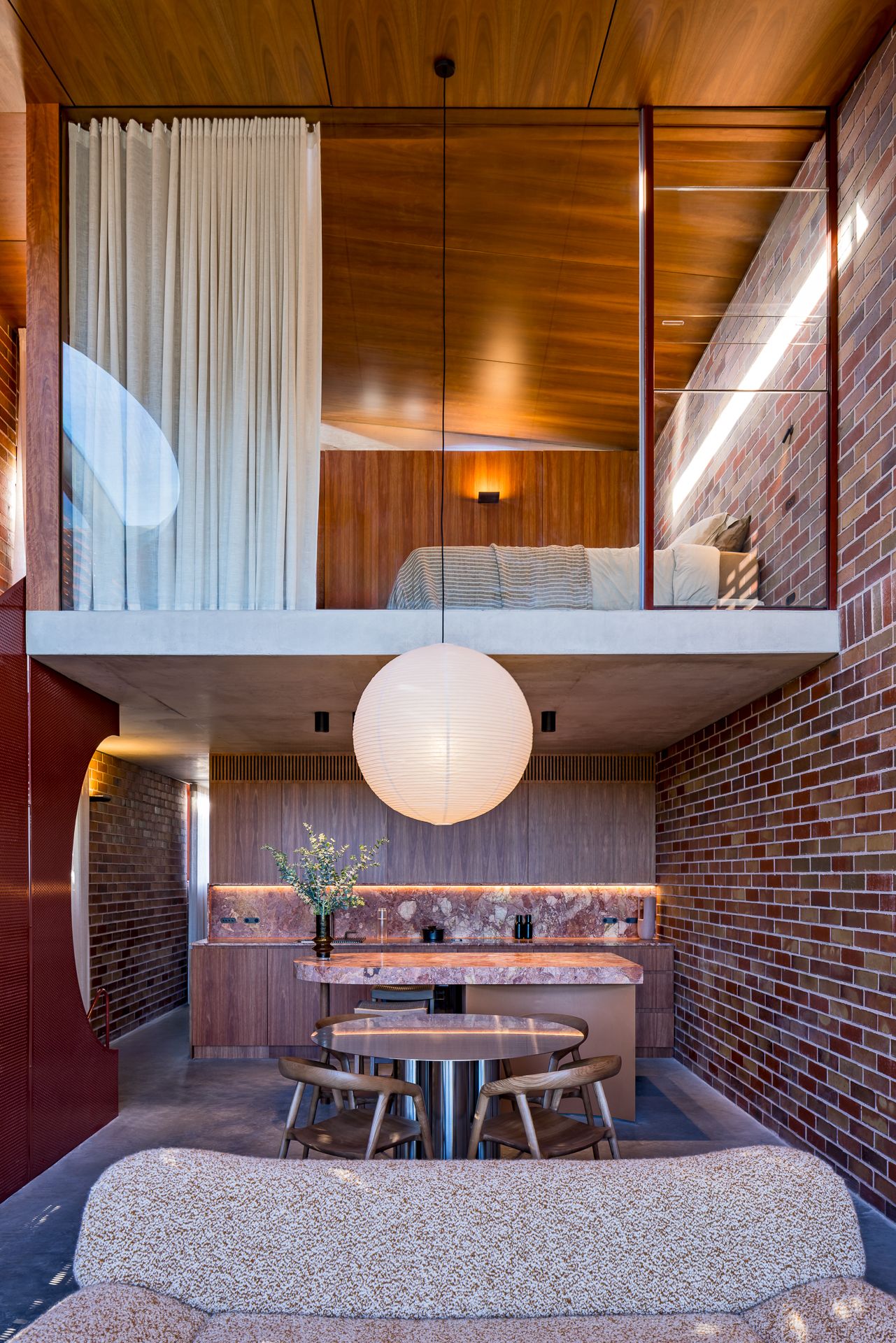 Double-height living space with exposed brick, timber-lined mezzanine bedroom, round paper pendant lamp and dining table below.