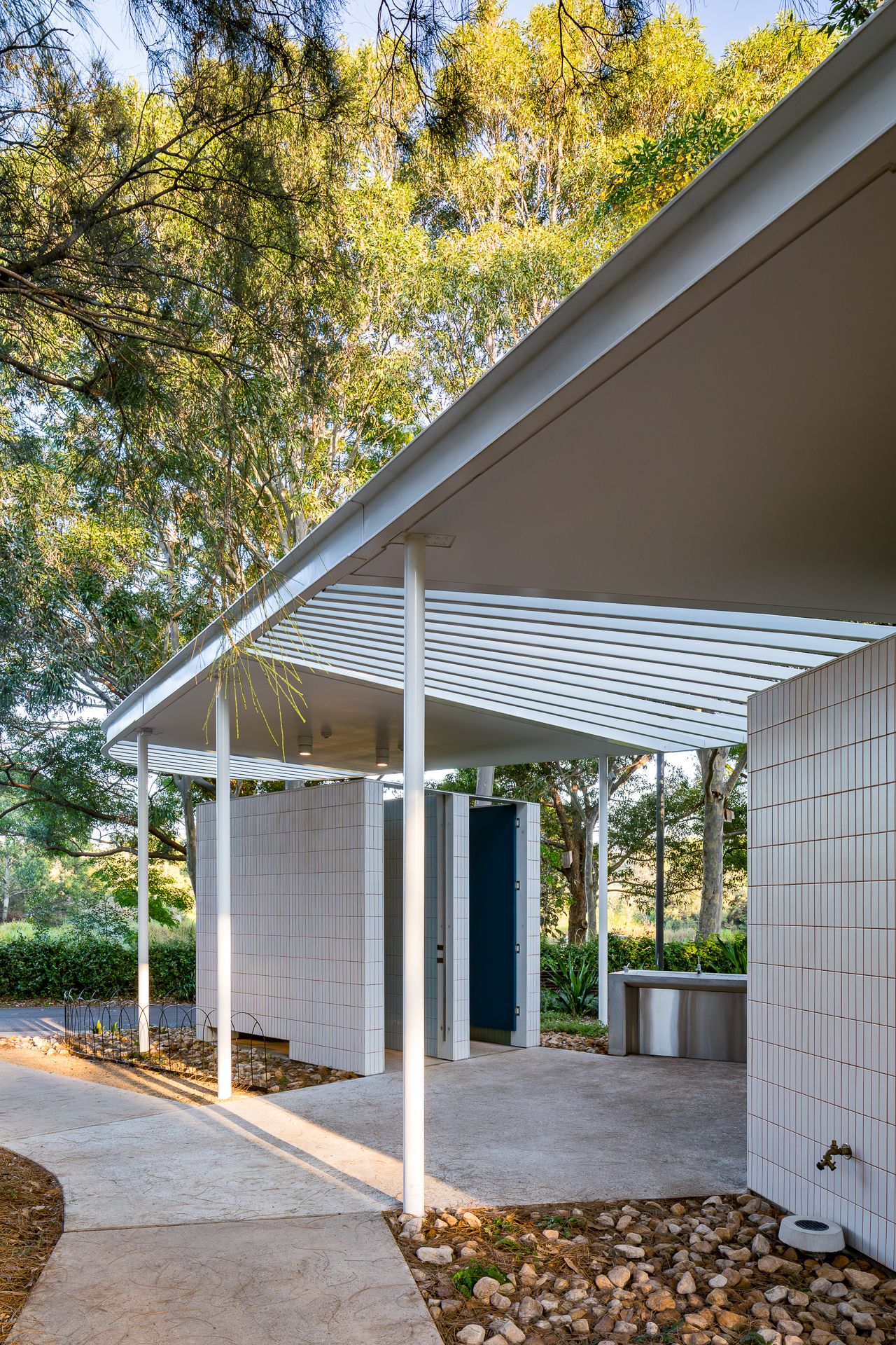 Covered walkway of a contemporary park restroom with white tiled walls, slim steel columns, a drinking fountain and river pebble garden bed.
