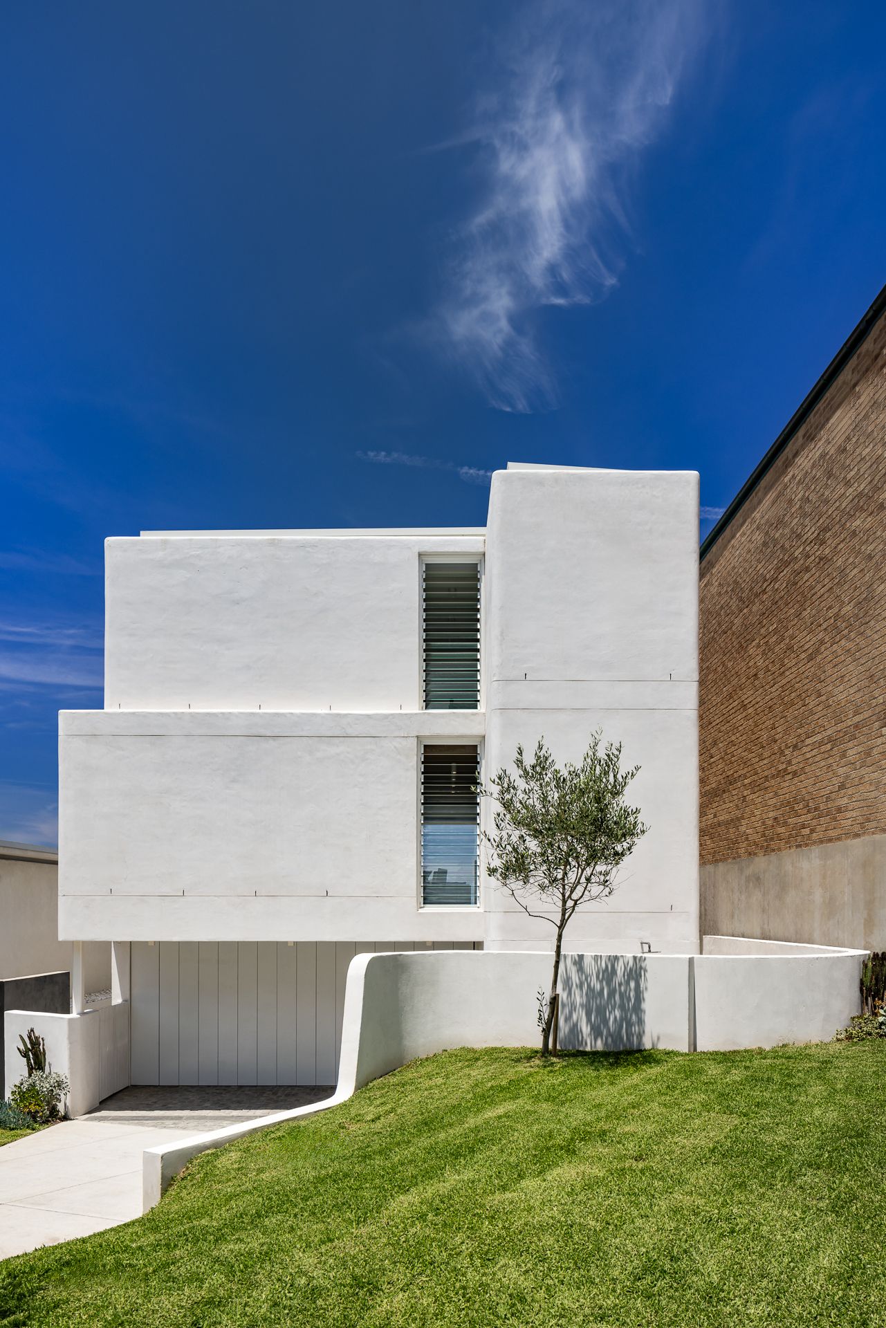 Modern white concrete house with louvred windows, an olive tree and a curved retaining wall above a green lawn under a blue sky.