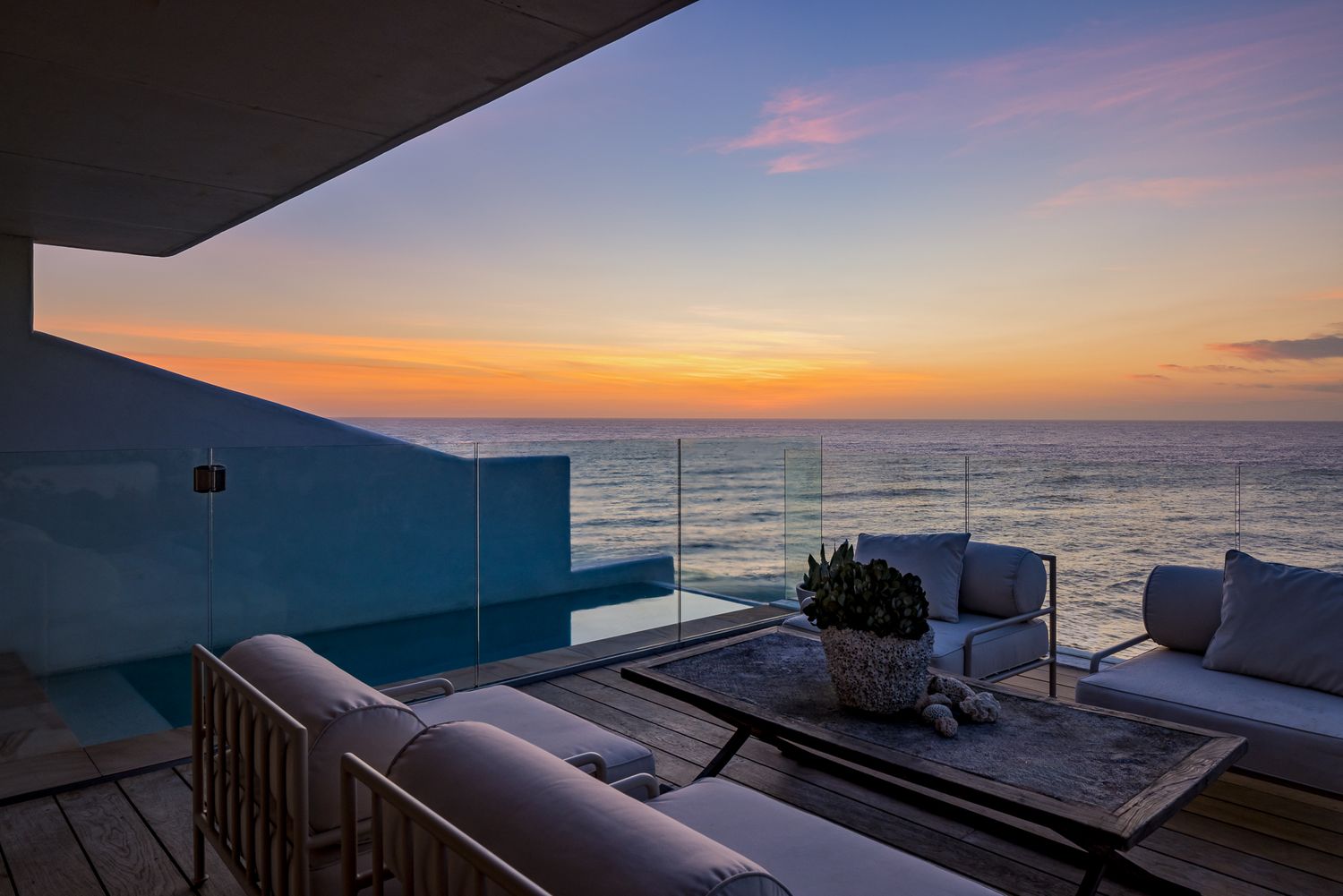 Oceanfront terrace with glass balustrade, outdoor sofas and a coffee table facing a vivid orange sunset over the sea.