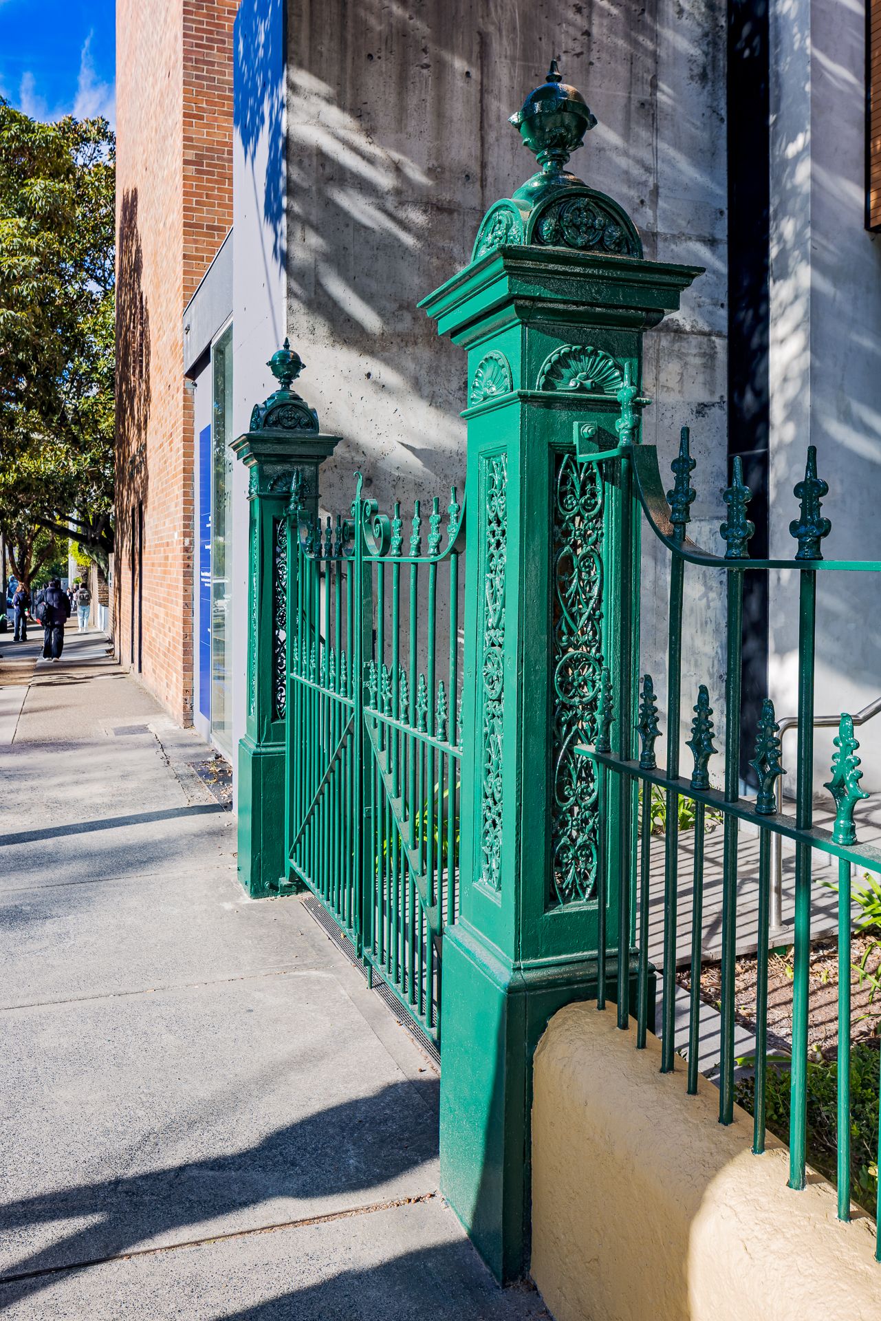 Ornate green cast iron gate and decorative pillars at a heritage building entrance on a sunlit city footpath.