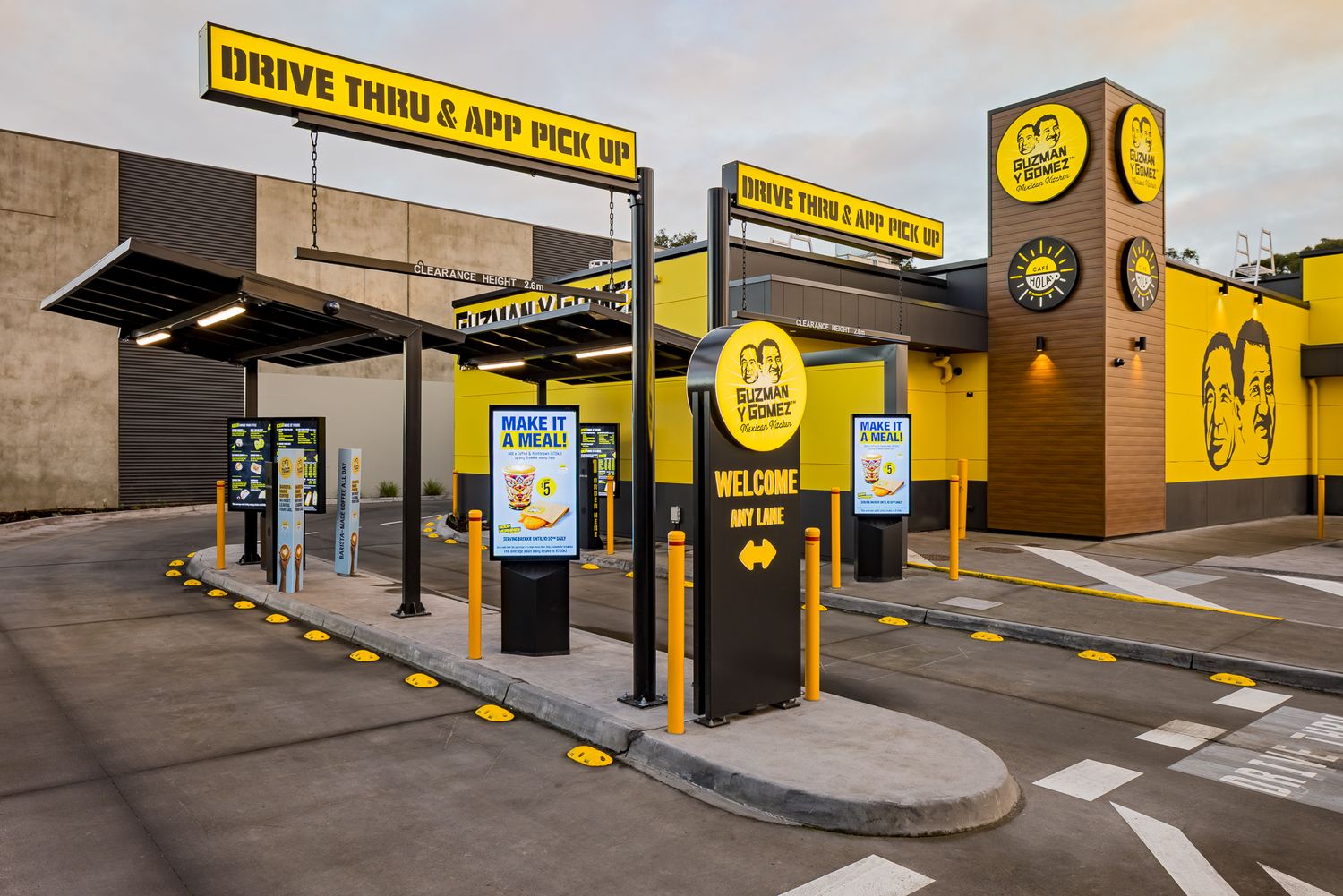 Guzman y Gomez drive-thru app pick up area with overhead signs, order screens and a yellow building with a branded brick tower.