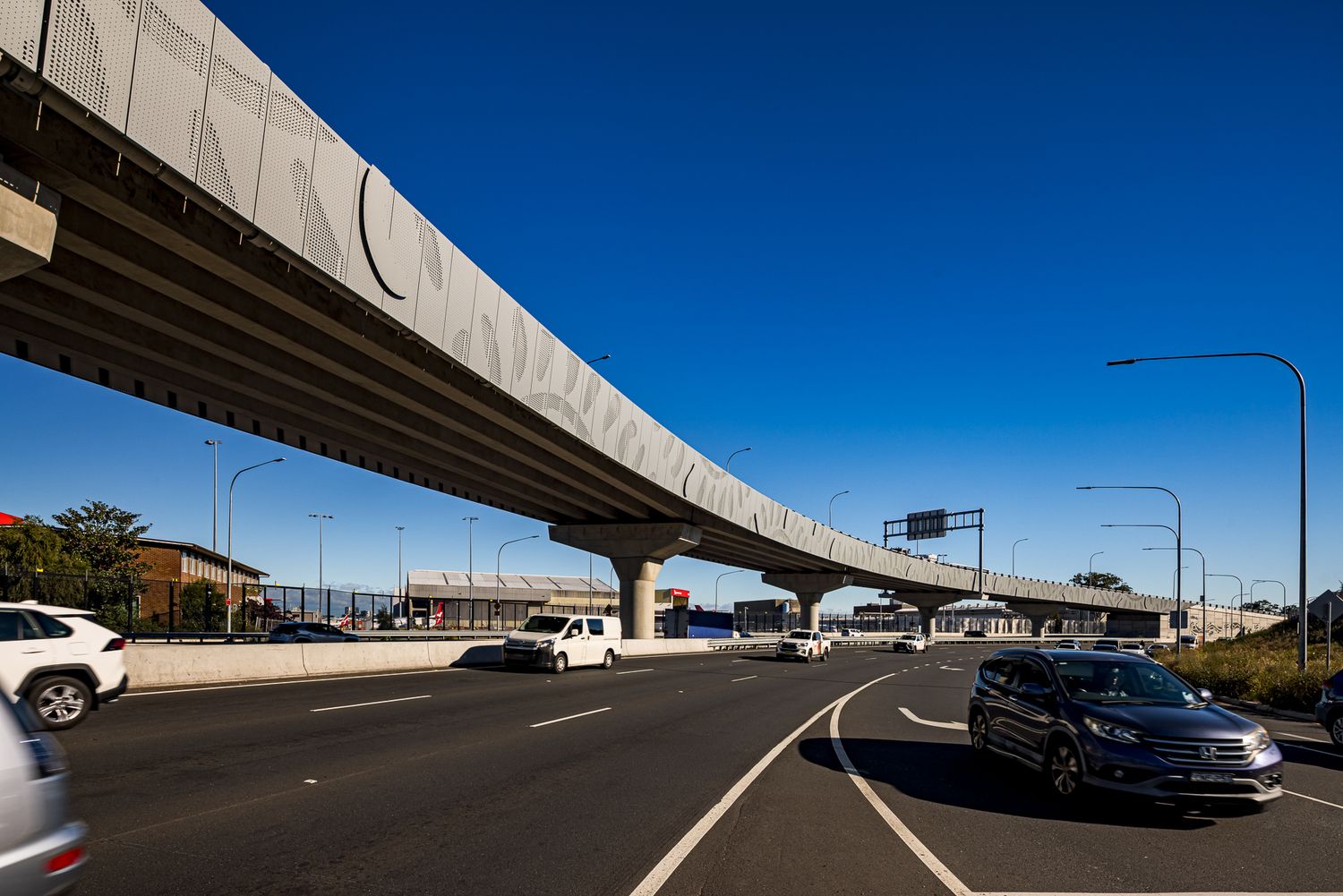 Elevated concrete motorway overpass with a decorative panelled barrier curving above multiple traffic lanes under a clear blue sky.