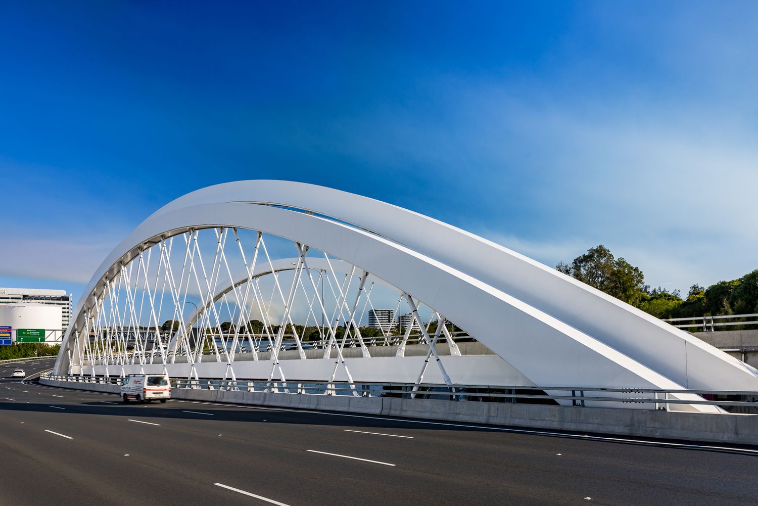 Curved white arch bridge with steel cable supports over a multi-lane highway, with a car driving beneath a deep blue sky.