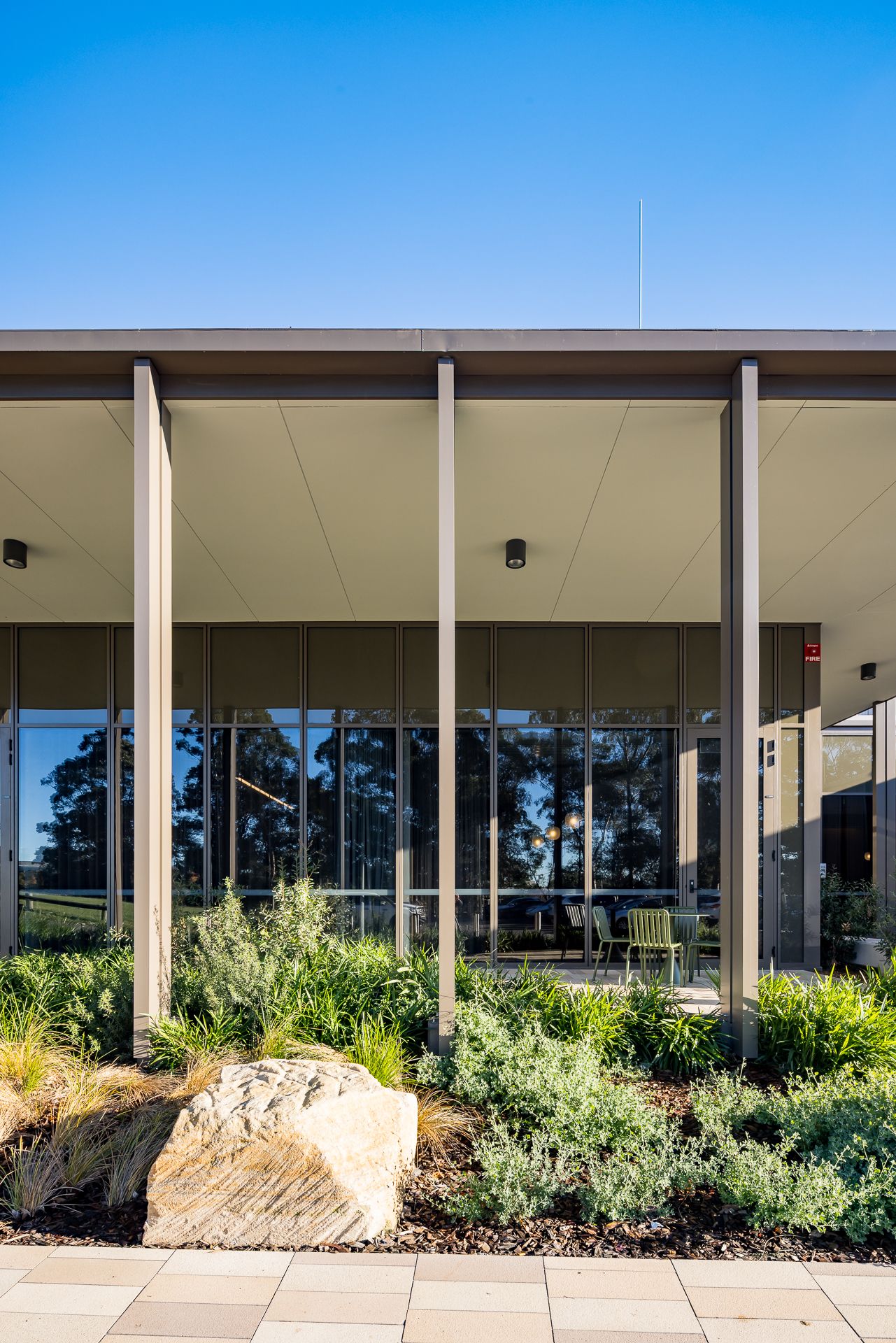 Covered walkway with slim steel columns and glass walls overlooks native shrubs and a large sandstone boulder under a blue sky.