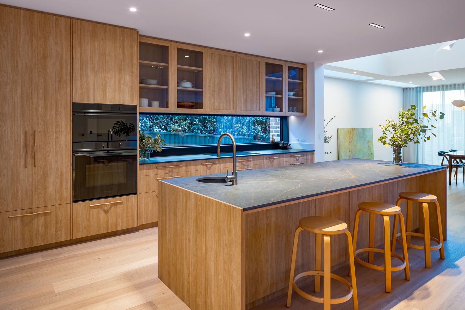 Warm timber kitchen with a stone-topped island, three wooden stools, black oven and a window splashback overlooking greenery.