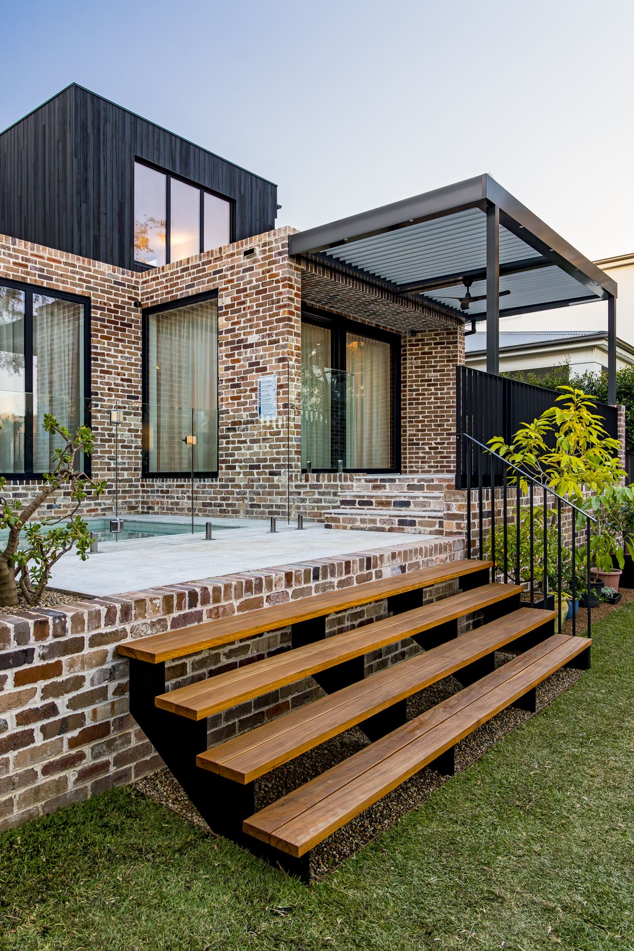 Brick and black timber home exterior with floating timber steps, a pool terrace, steel pergola and landscaped garden at dusk.
