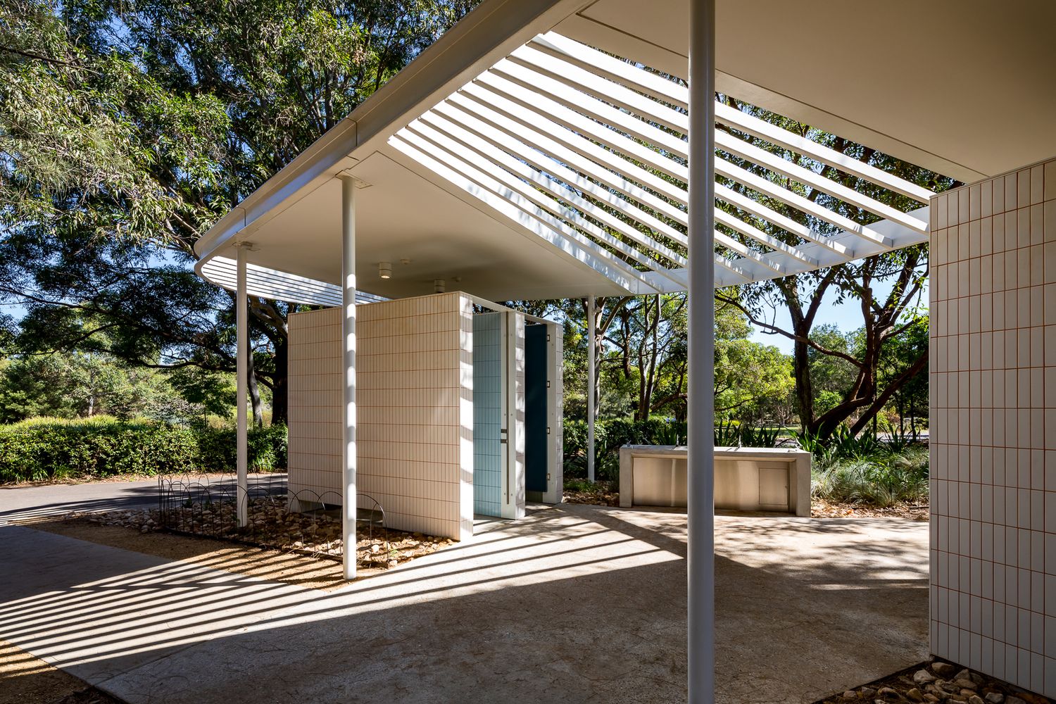 Tiled public amenities building with slim steel columns and a slatted roof casting striped shadows on the paved path.