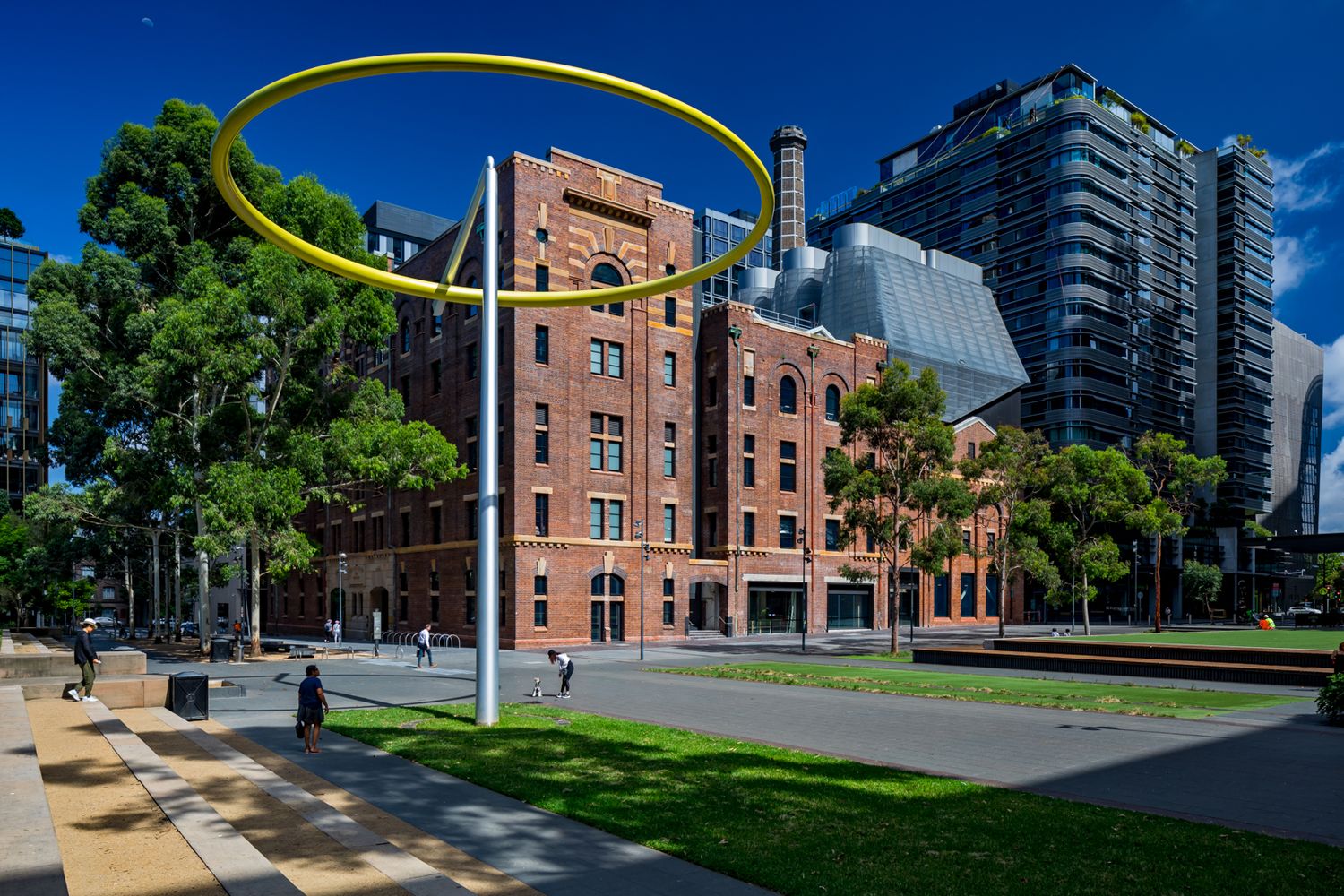 Historic red brick building beside modern towers with a giant yellow ring sculpture rising above a landscaped urban plaza.