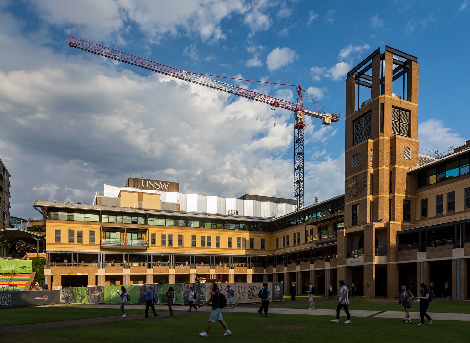 University campus building with UNSW sign, brick tower and construction crane as students play on the sunny lawn.