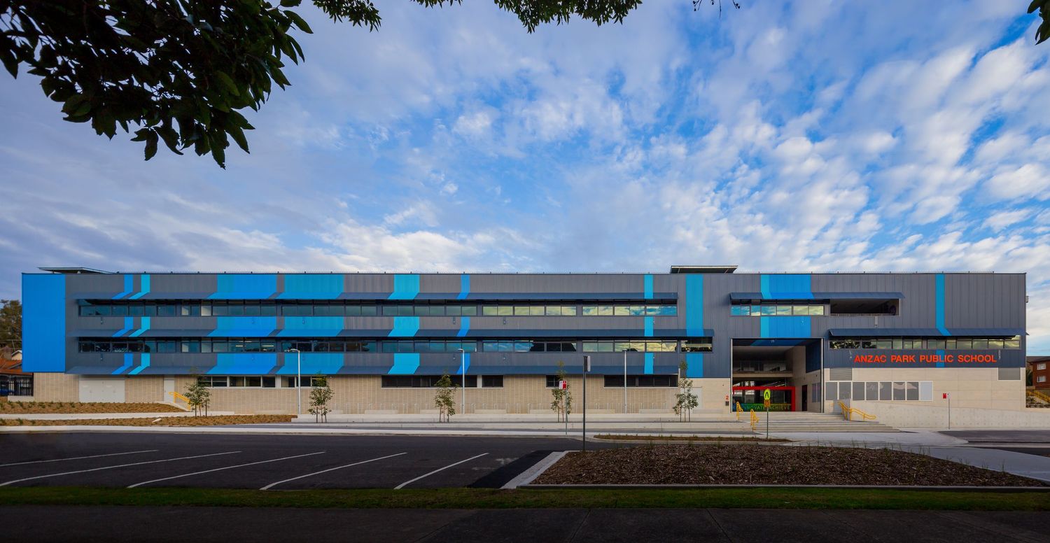 Long modern two-storey building with blue and grey cladding and a car park under a blue sky with scattered clouds.