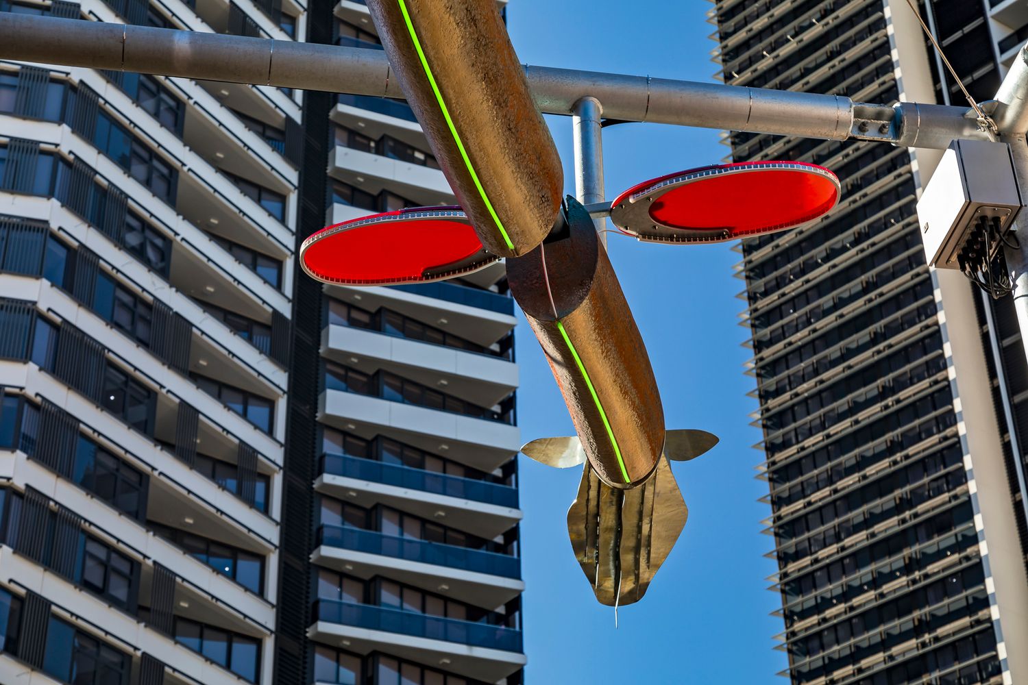Fish-shaped suspended sculpture with red oval discs hangs above a city street between tall apartment towers under a blue sky.