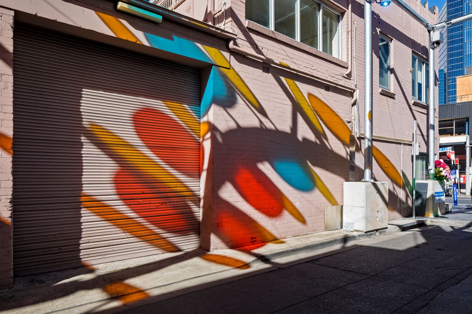 Narrow city alley with a pink brick wall and roller door painted in bright red, blue and yellow shapes under strong sunlight.