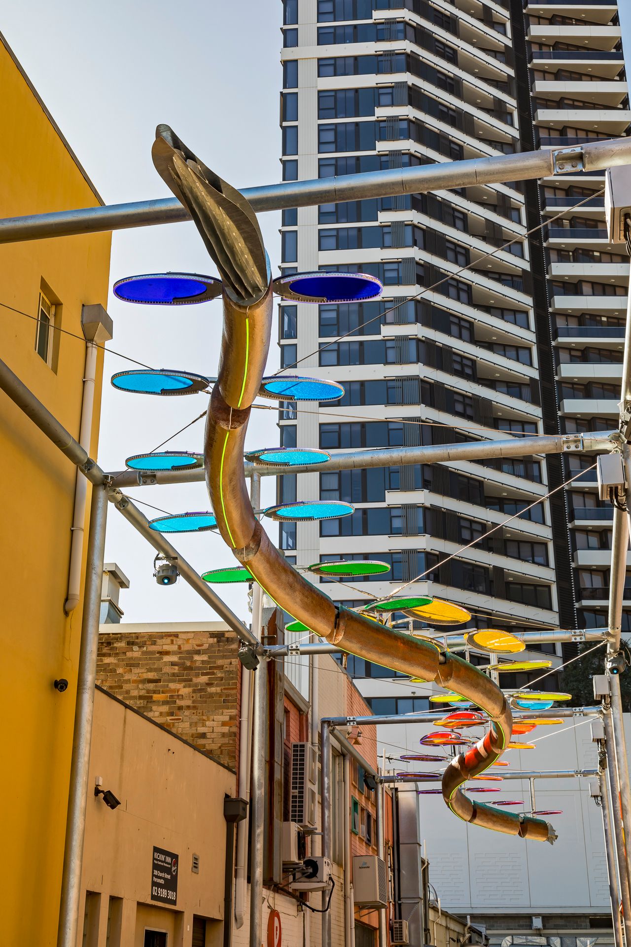 Serpentine metal sculpture with blue, green and orange discs suspended above an urban laneway beside a yellow building and tower.