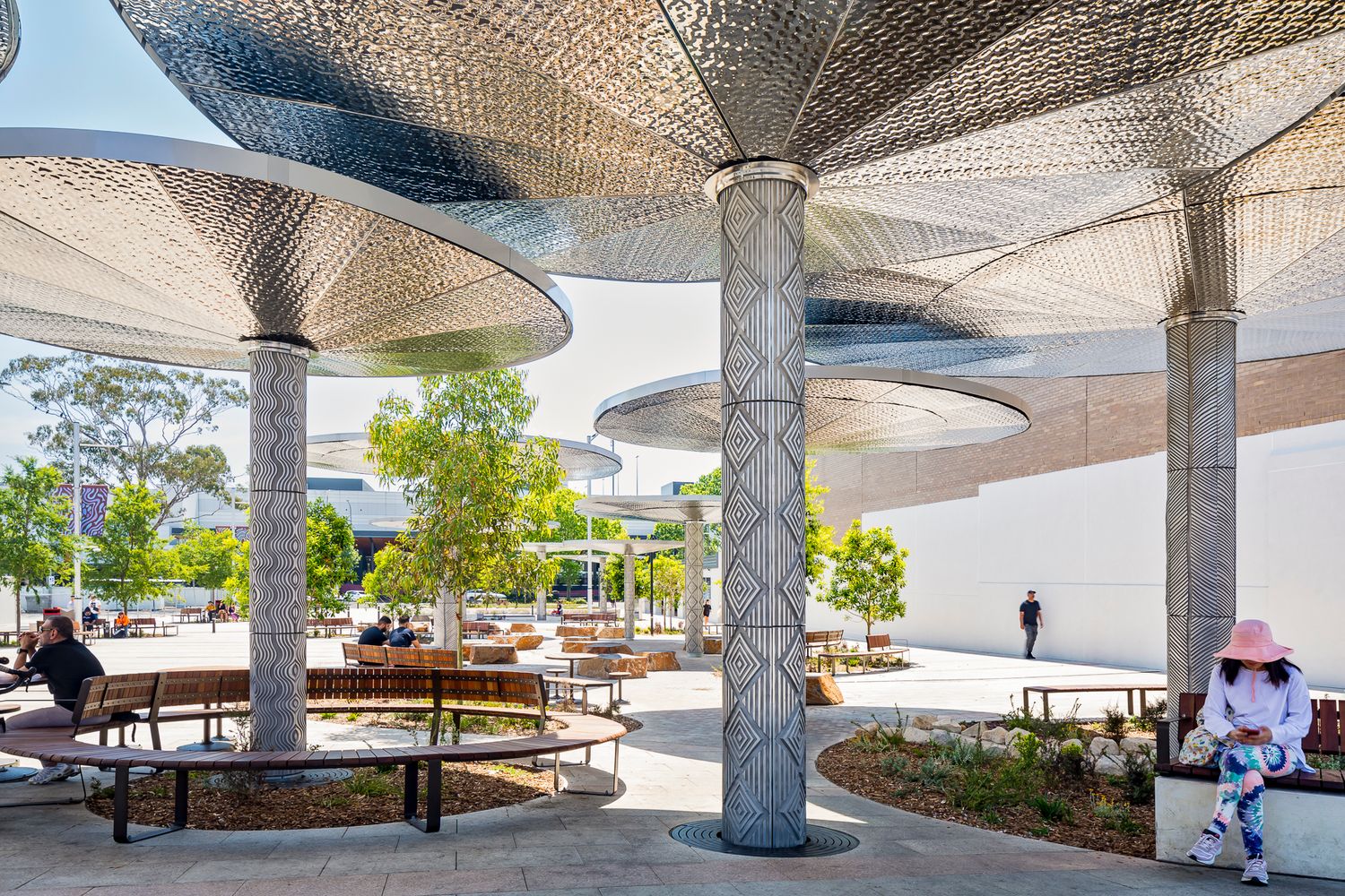 Textured metal columns and canopy shades over wooden benches and planted beds in a bright urban plaza.