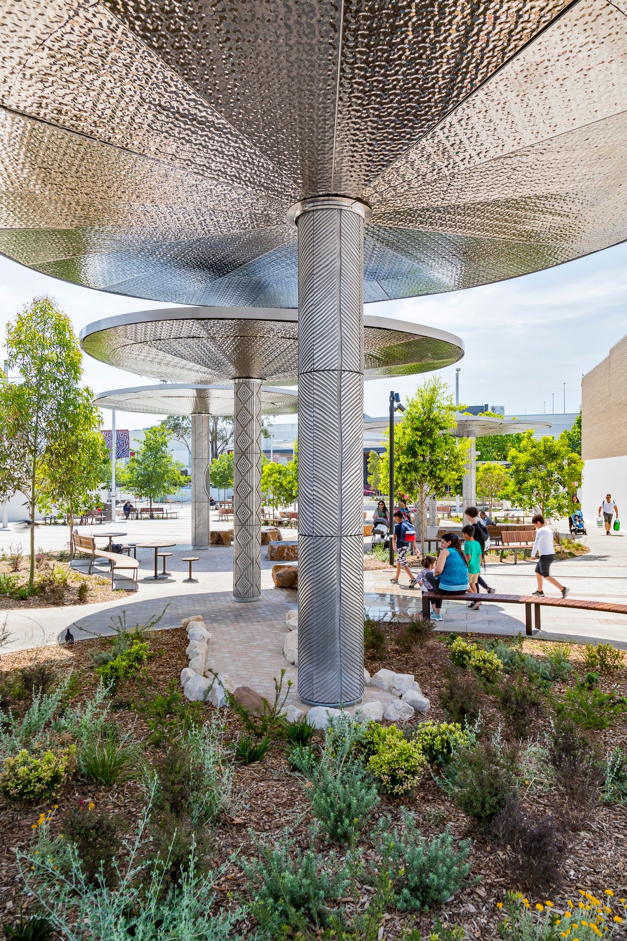 A textured metal column supports a large circular canopy above native gardens, benches and visitors enjoying a vibrant public plaza.