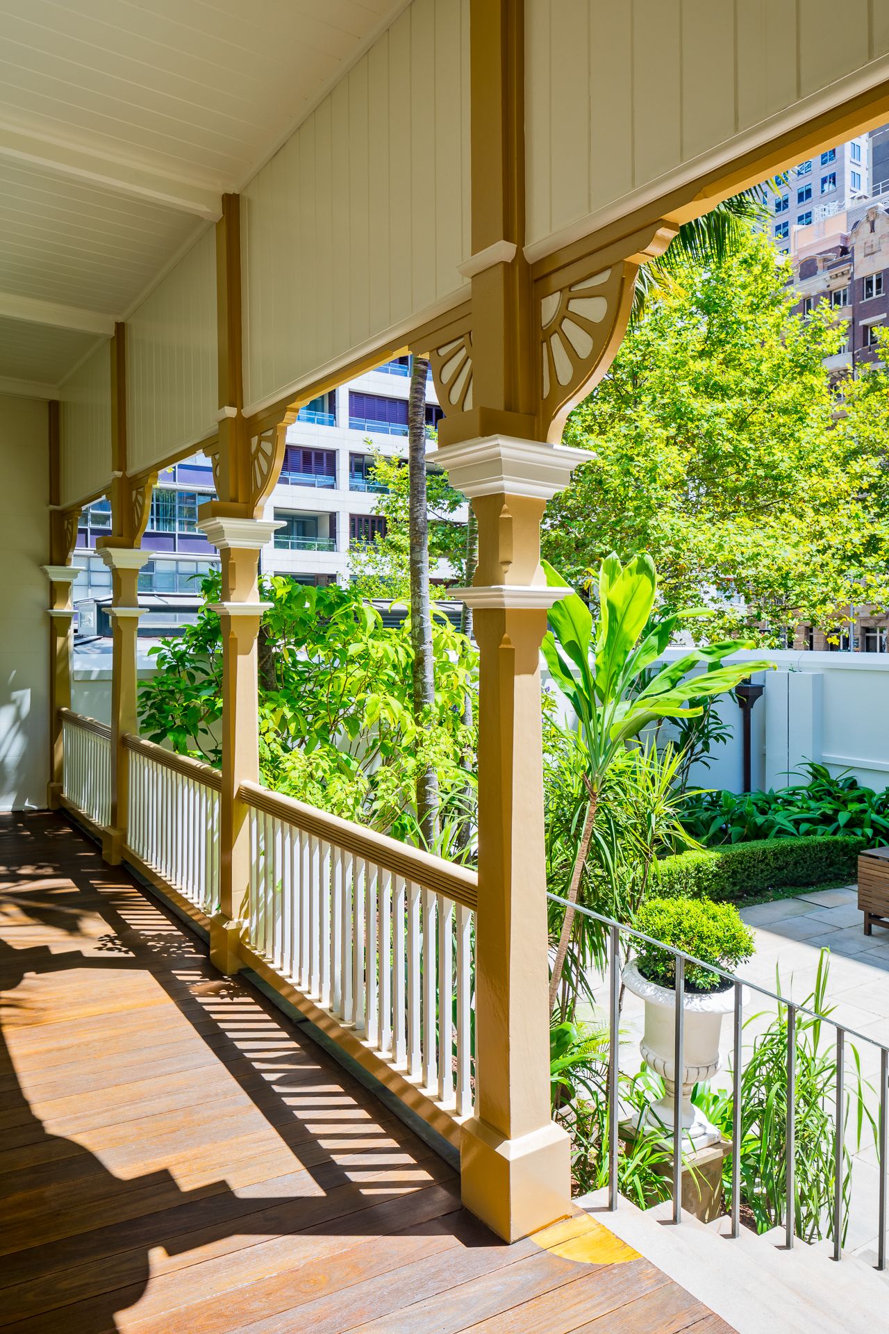 Sunlit timber veranda with white railings and decorative brackets overlooking lush tropical gardens and city buildings.