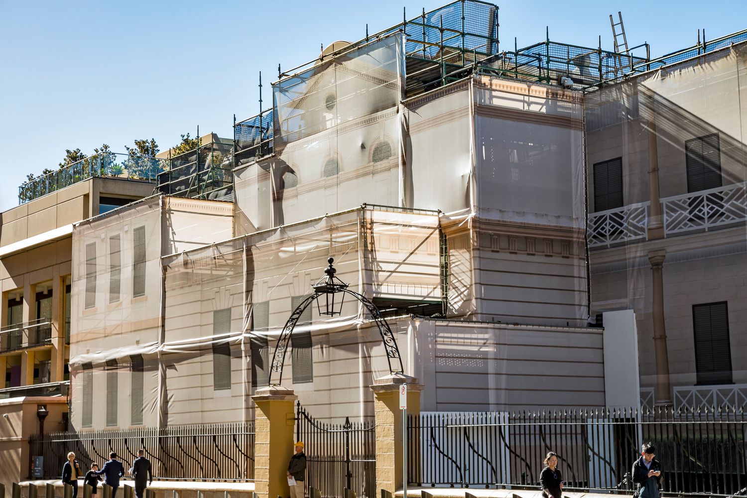 Historic building corner enclosed in protective sheeting and scaffolding during restoration, with pedestrians walking by the iron fence.