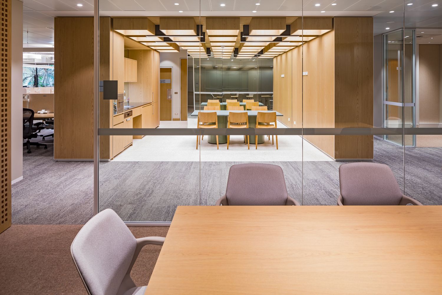 View through glass partitions of a wood-paneled office meeting room with a conference table, chairs and a kitchen area beyond.