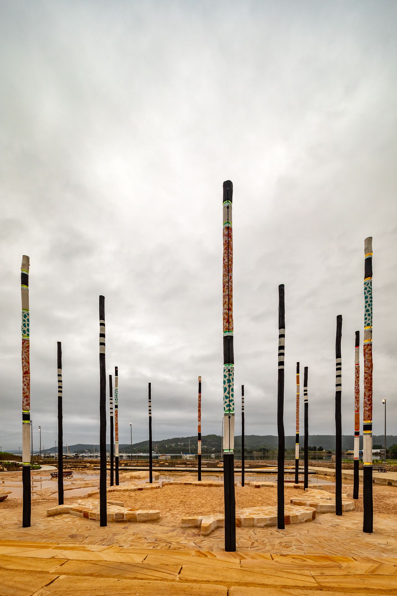 A circle of tall painted Aboriginal art poles stands on a sandstone plaza with hills and cloudy sky in the background.