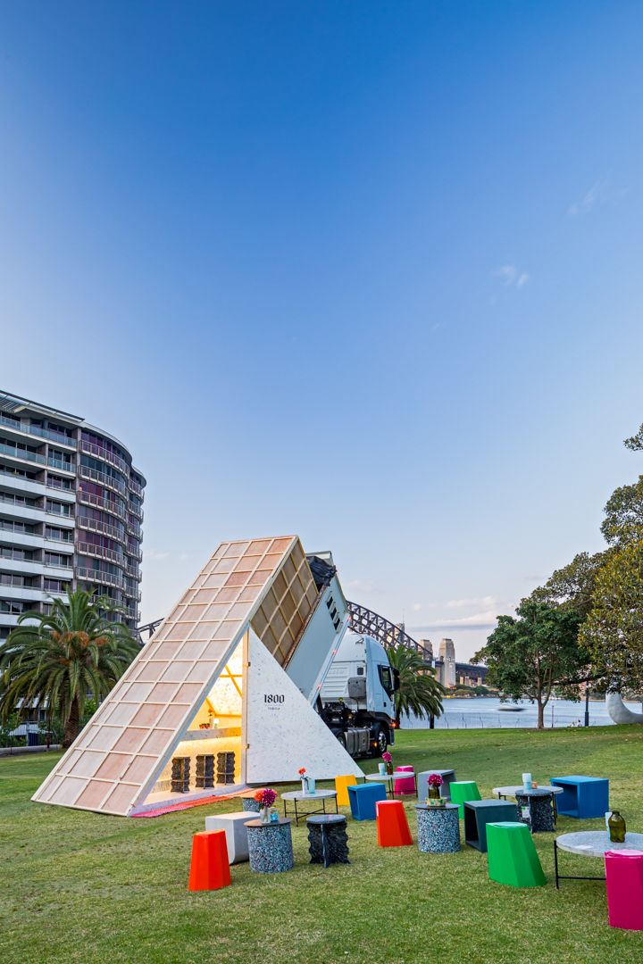 Glowing triangular pop-up pavilion on a waterfront lawn with colorful stools and tables, a truck, palm trees and apartment buildings under b