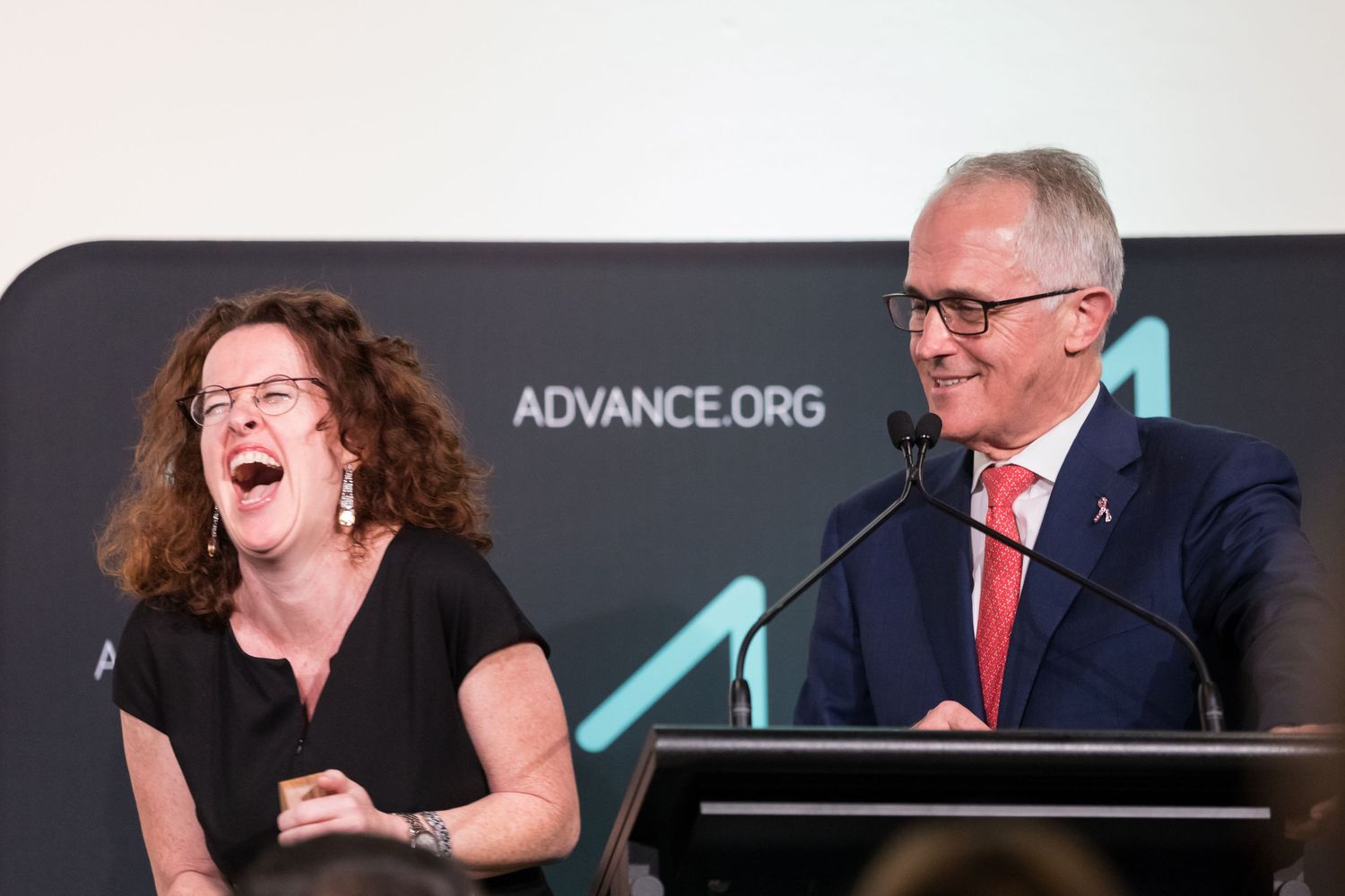 A woman laughs heartily beside a smiling man in a suit and glasses speaking at a podium with microphones at an Advance.org event.