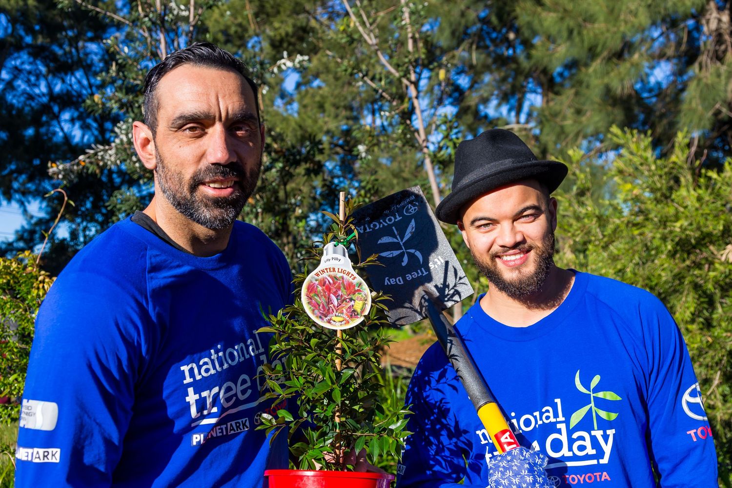 Two smiling men in blue National Tree Day shirts hold a potted plant and a small shovel outdoors among green trees.