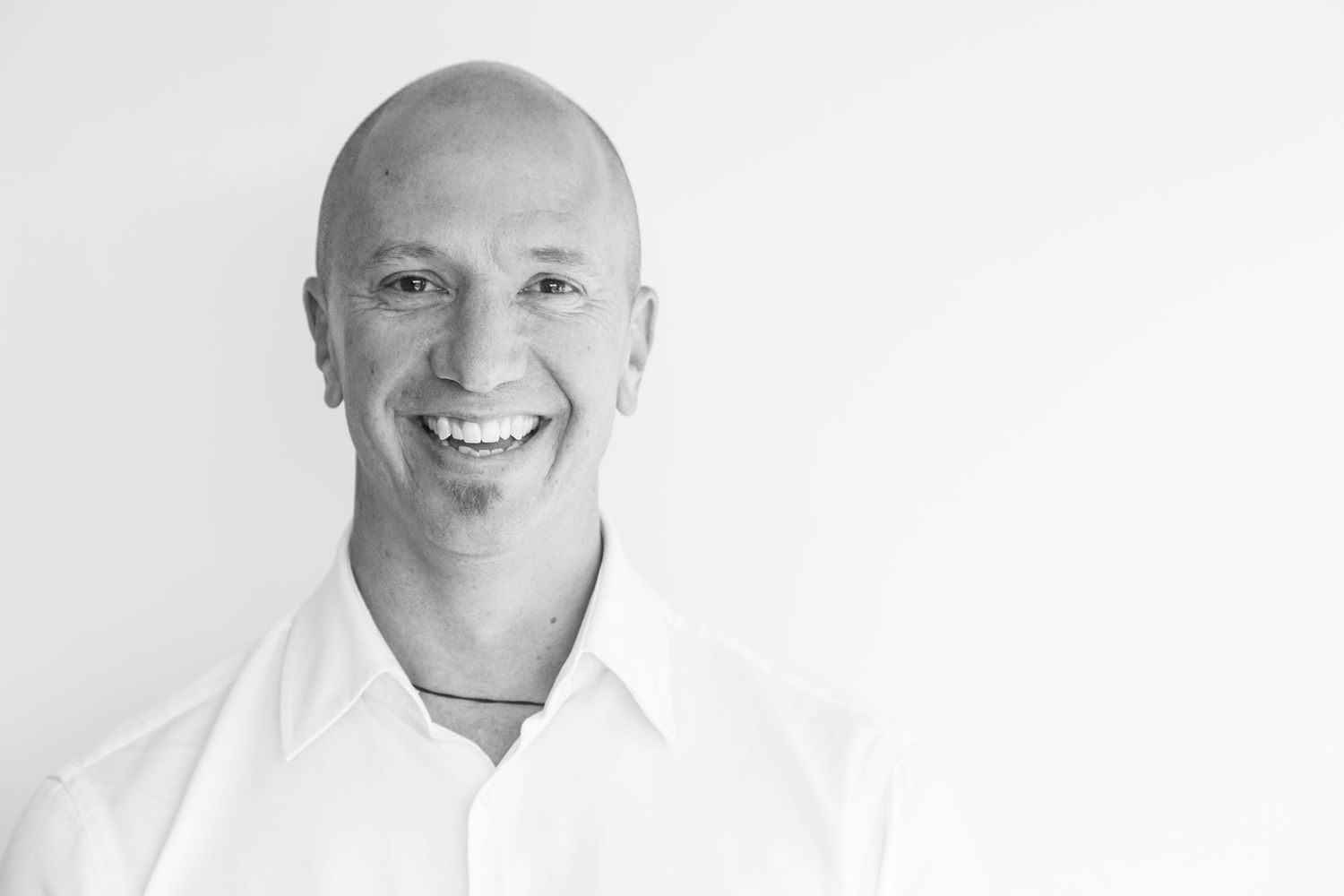 A black and white portrait of a smiling bald man in a white collared shirt against a plain light background.